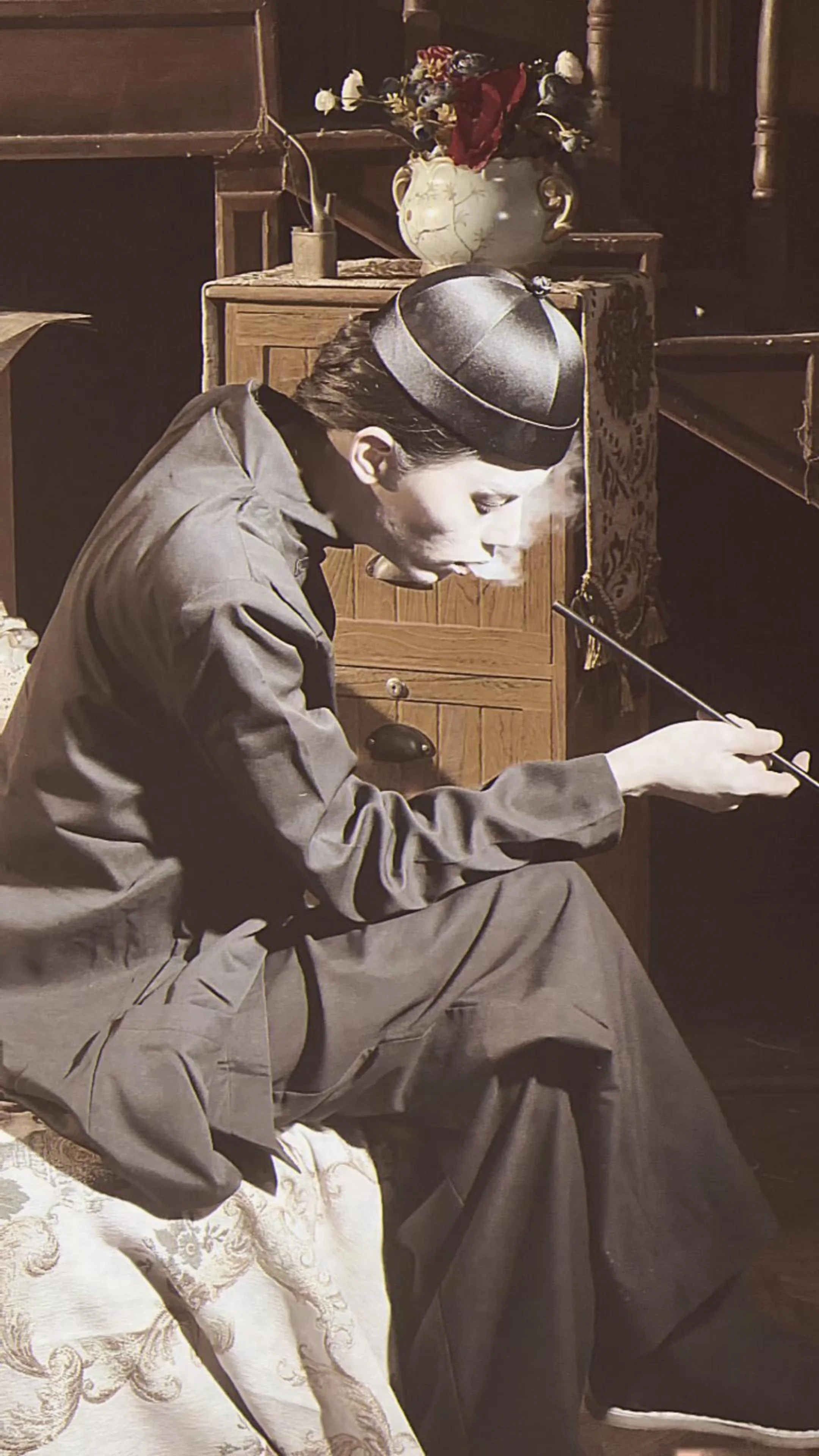 A black and white photo of a man sitting on a bed, smoking a cigarette, wearing a cap and a long coat.