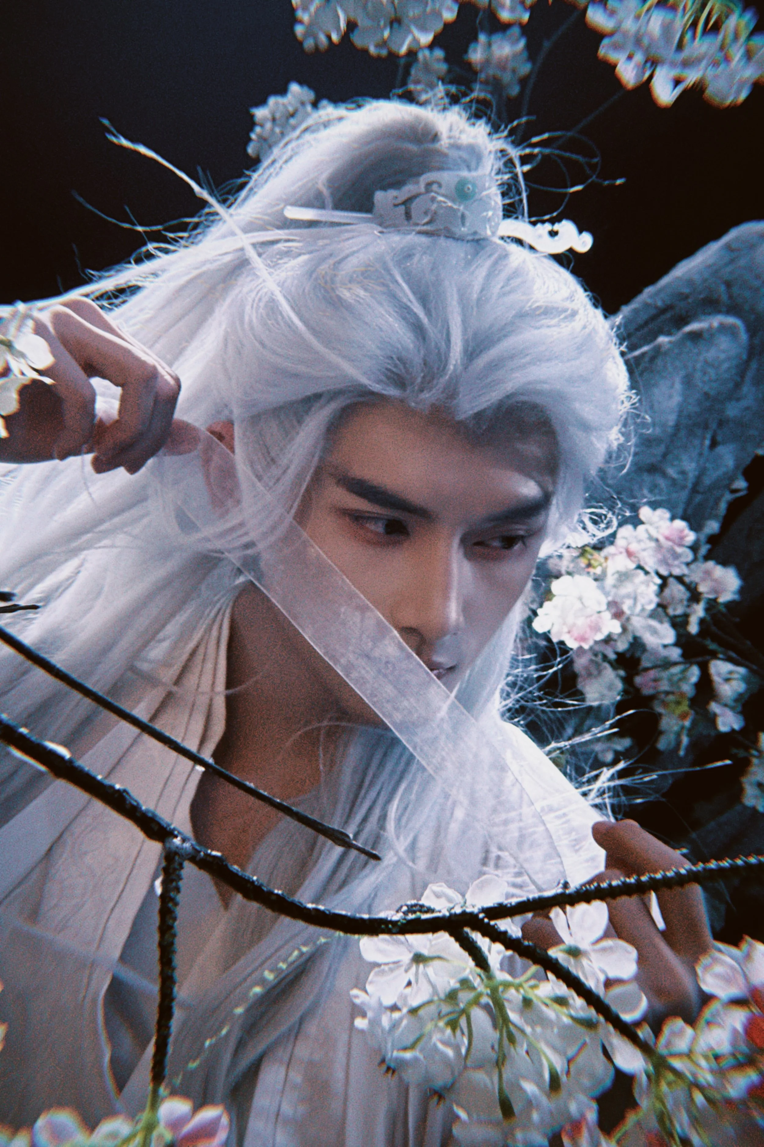 A person with long, white hair, wearing white clothing, surrounded by flowers and branches.