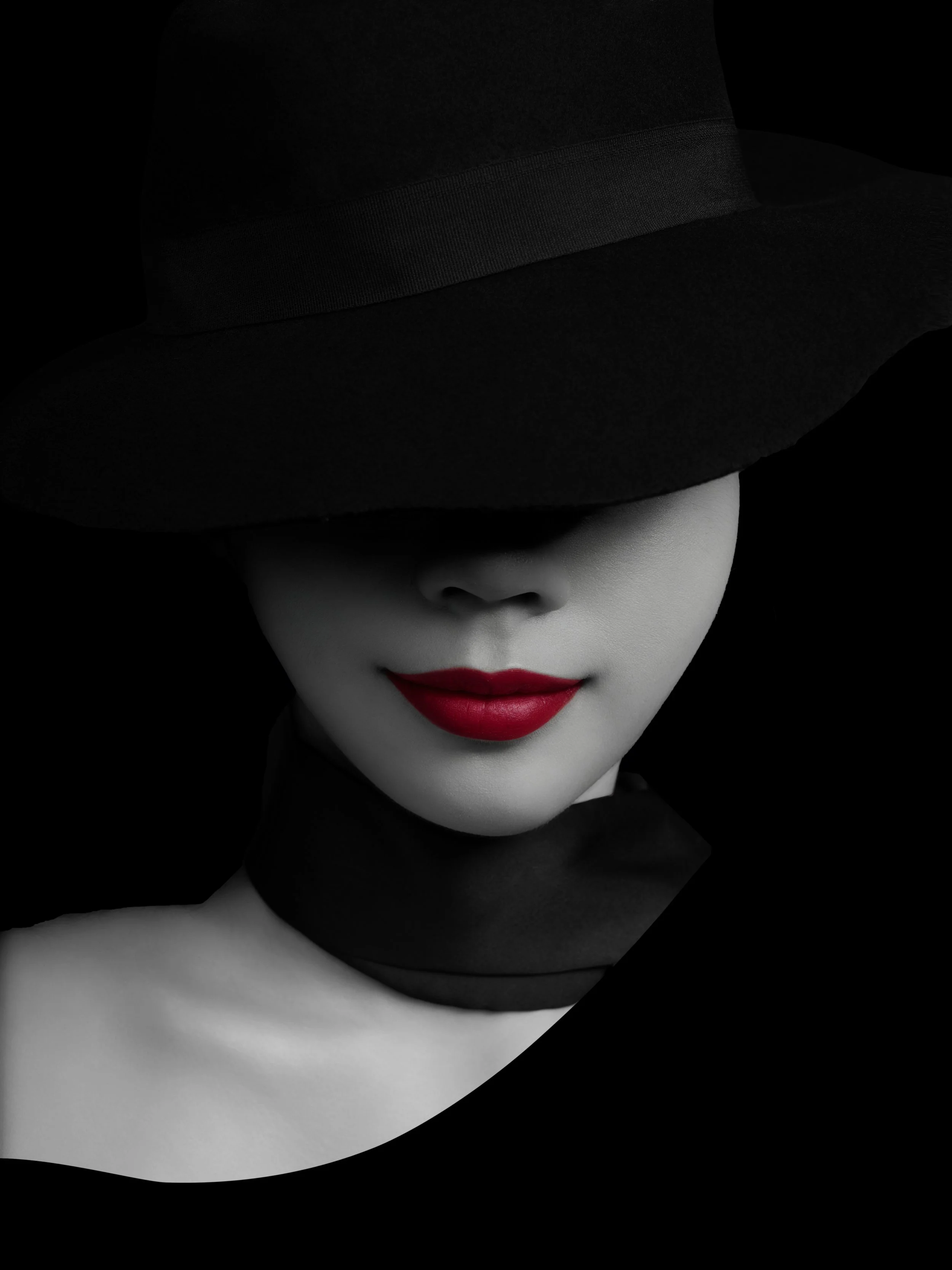 A woman with painted lips wearing a large black hat that covers her eyes, with a black choker and a black strapless top, against a black background.