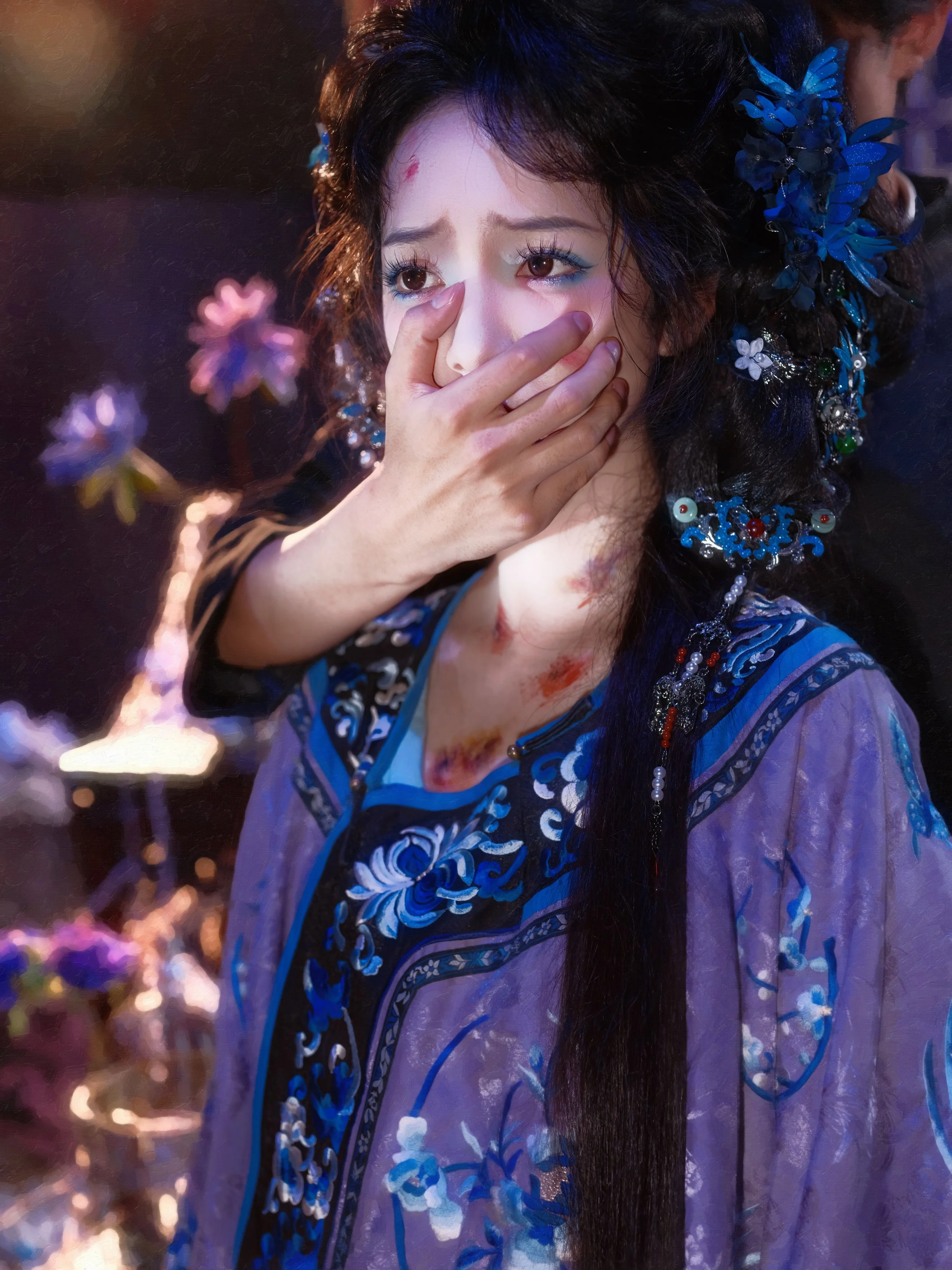 A woman in traditional Asian clothing with intricate blue embroidery and accessories, covering her mouth with her hand. She appears to be distressed or emotional.