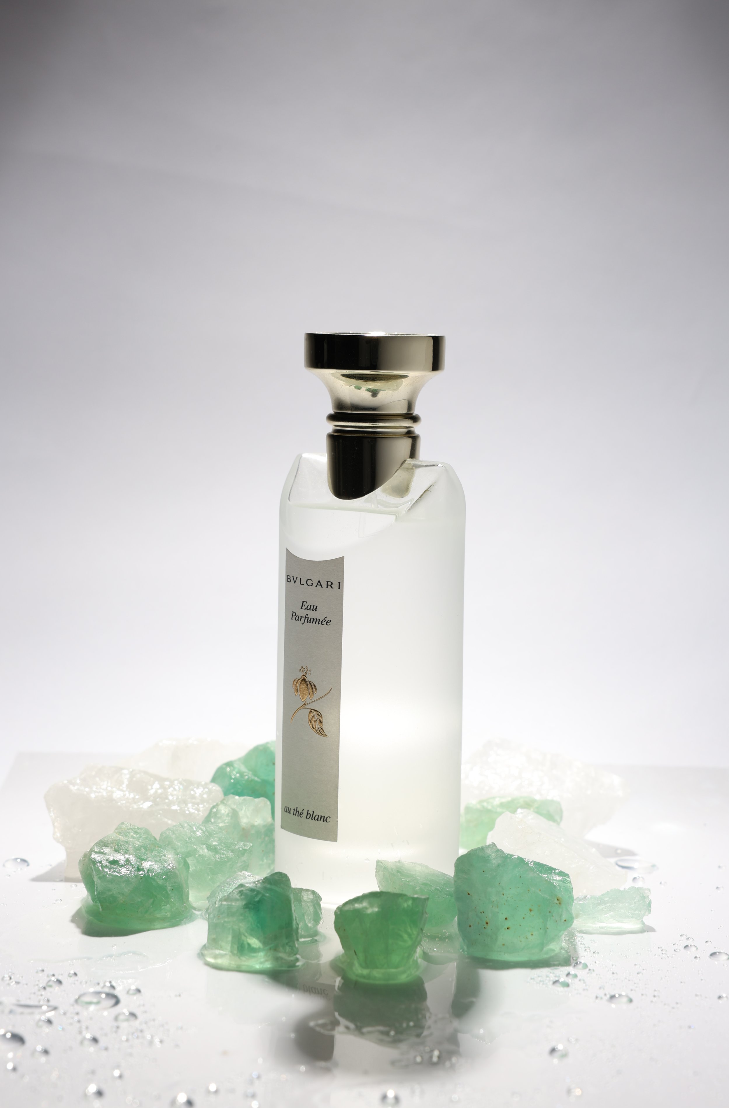 A fragrance bottle surrounded by green and white mineral rocks on a white surface with water droplets, with a plain gradient background.
