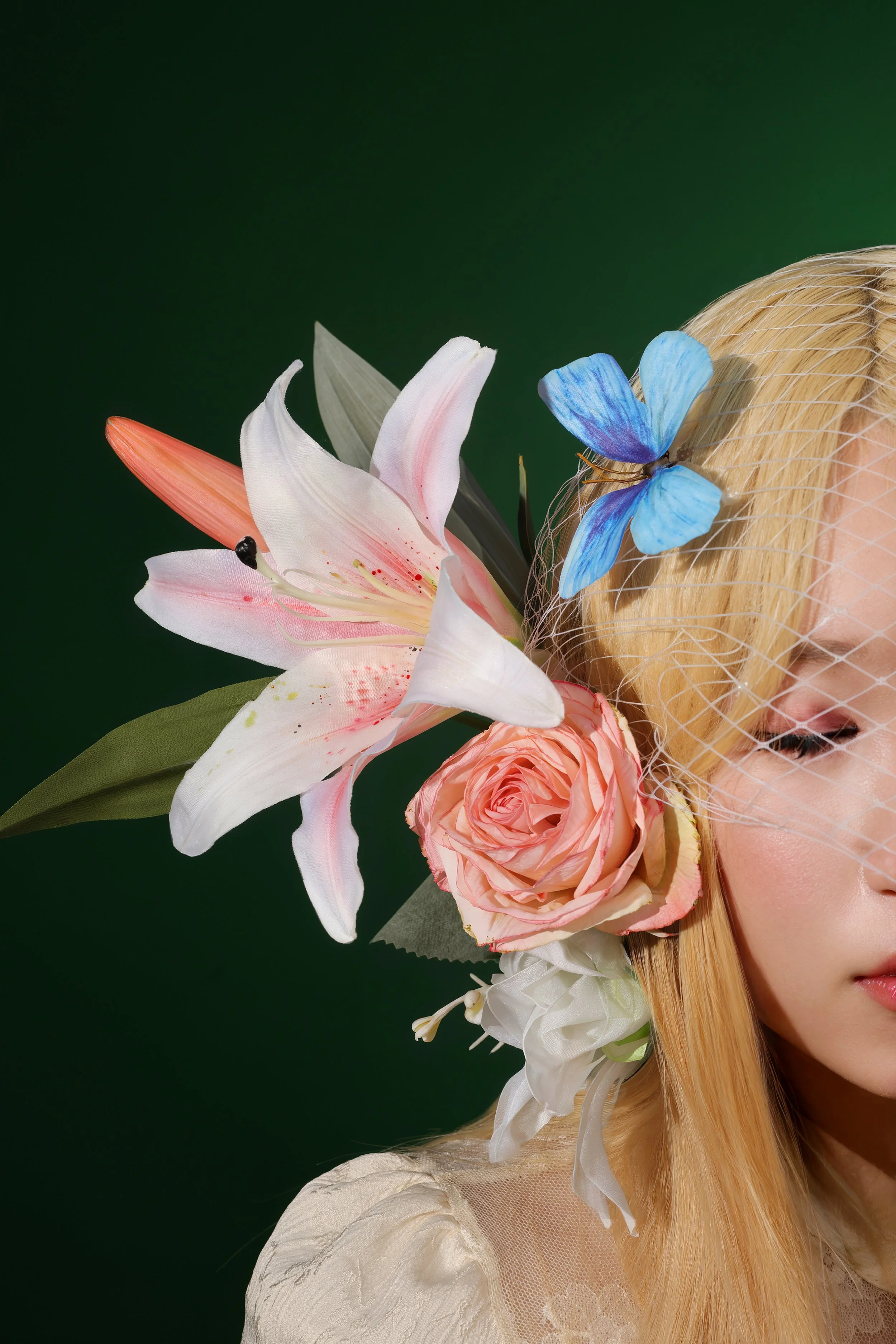 A woman with blonde hair features floral accessories including a pink lily, a pink rose, and a blue butterfly on her head against a green background.