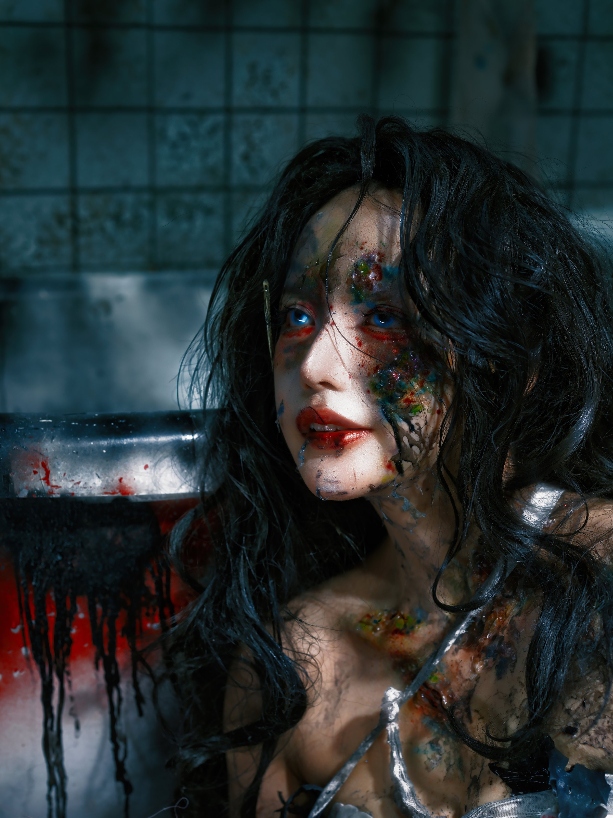 A woman with black, messy hair and blue eyes, covered in colorful paint and blood, with a sinister expression, in a dim, tiled room.