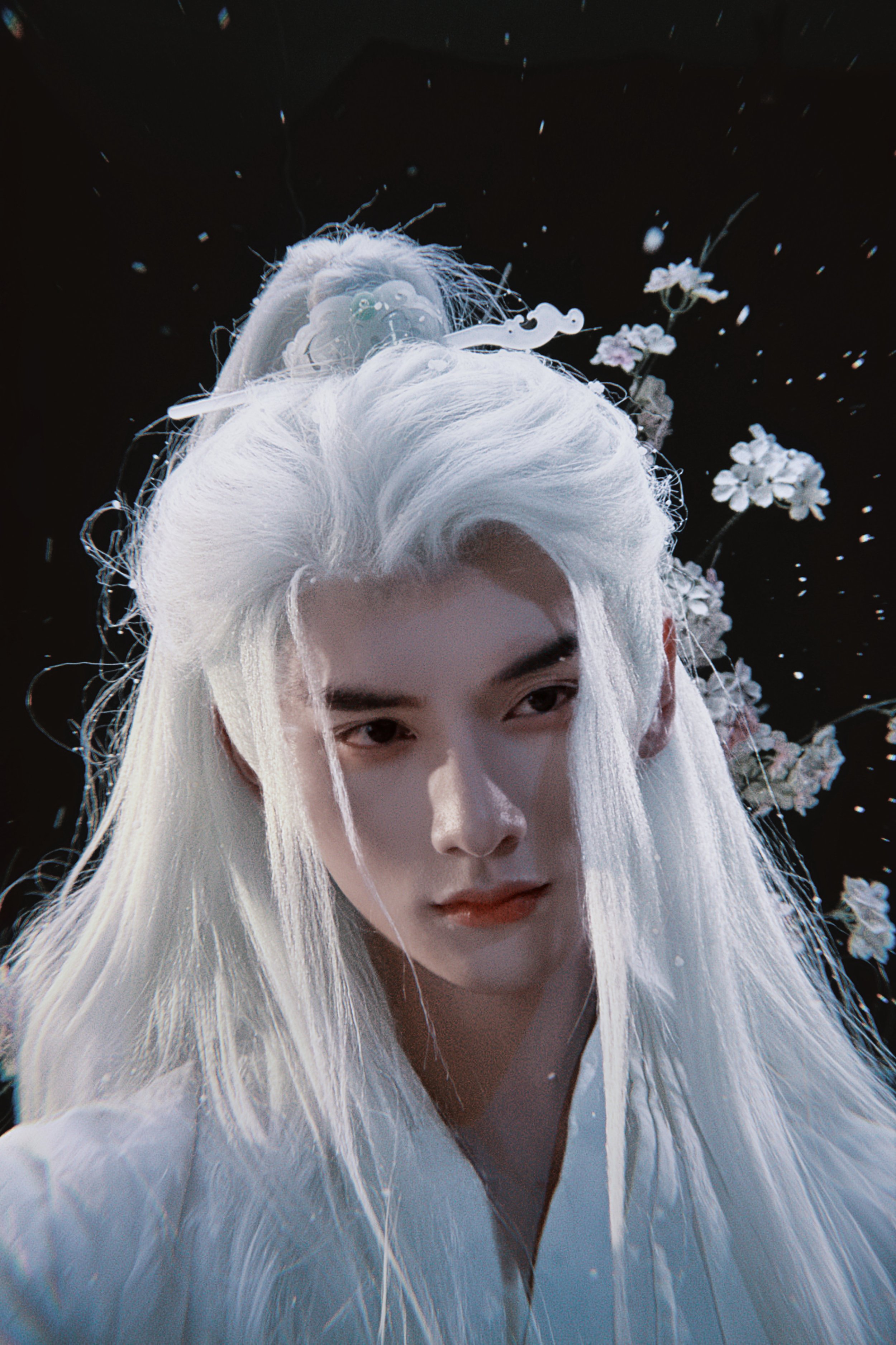 A person with long, white hair styled in an elaborate fashion, wearing traditional clothing, with an ornate headpiece adorned with flowers and decorations, surrounded by falling flower petals against a dark background.