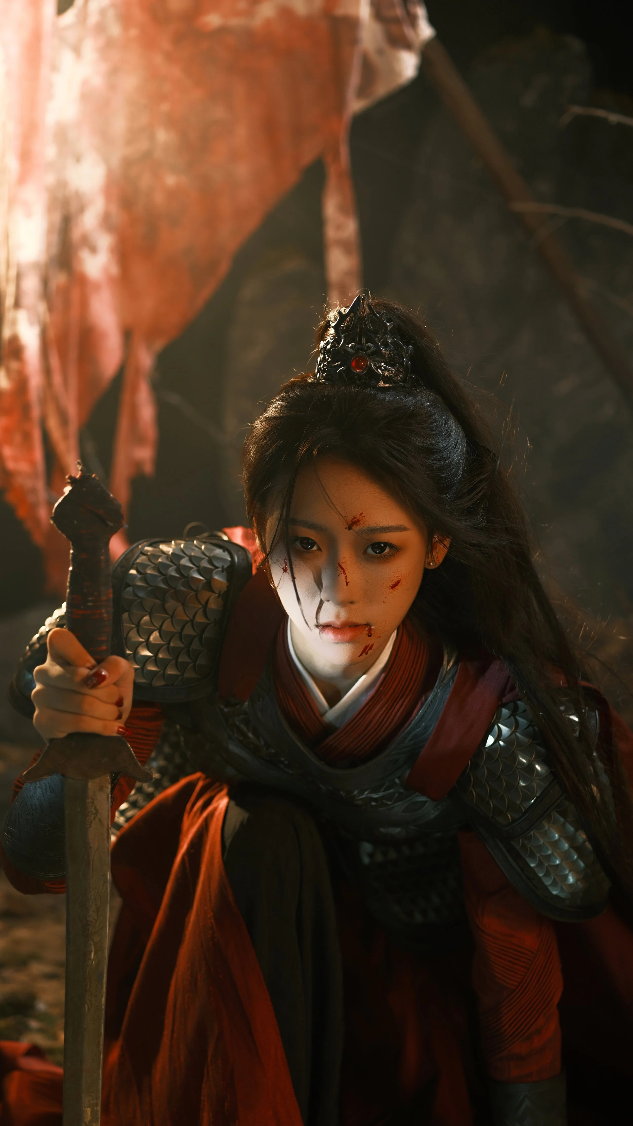 A woman dressed as a fierce warrior in armor, with long black hair, holding a sword, and covered in dirt and blood, in a dramatic scene.