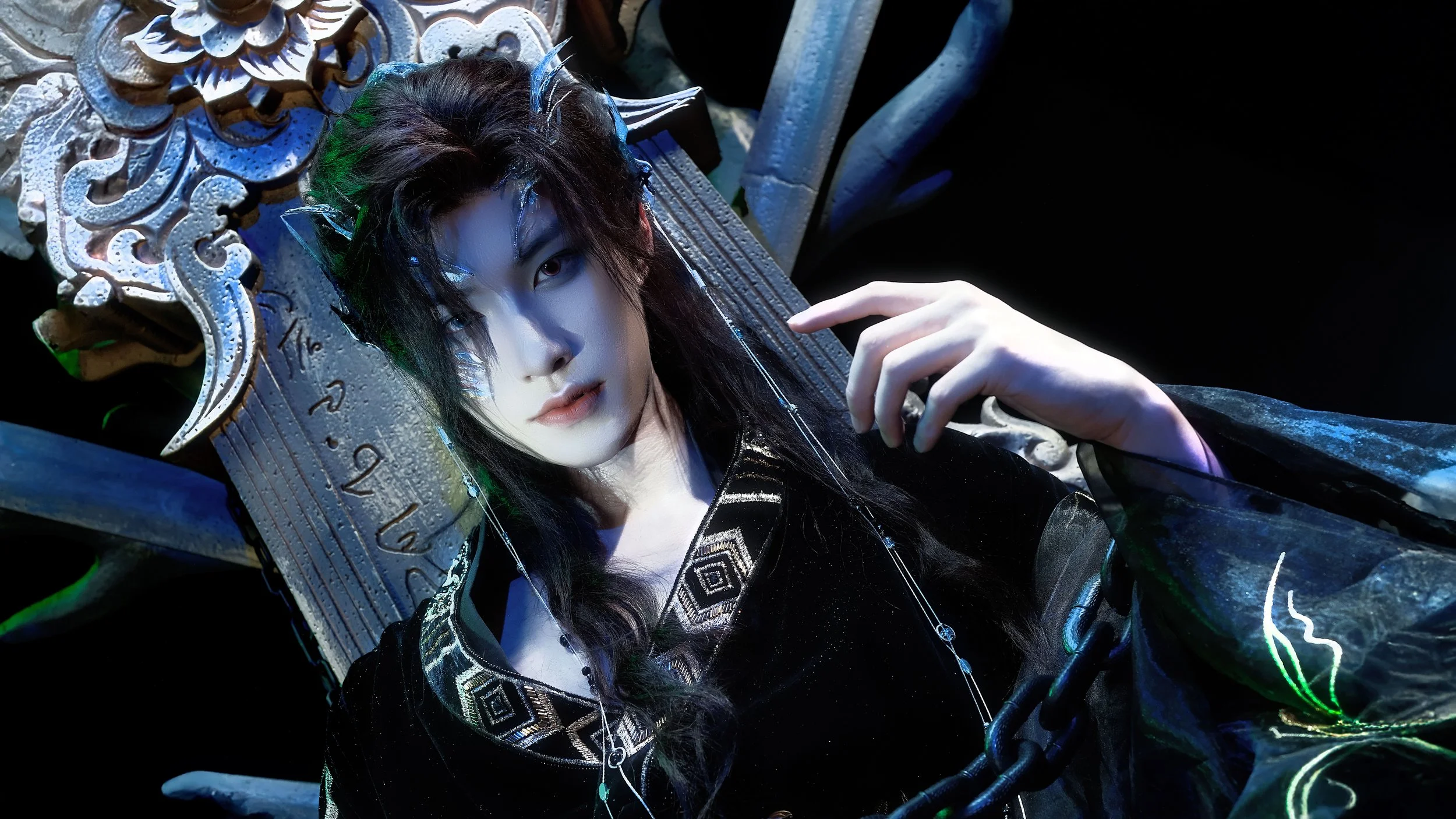 A person with elaborate makeup and costume, sitting on an ornate throne-like chair, with dark hair, intense gaze, and intricate details on their clothing and accessories.