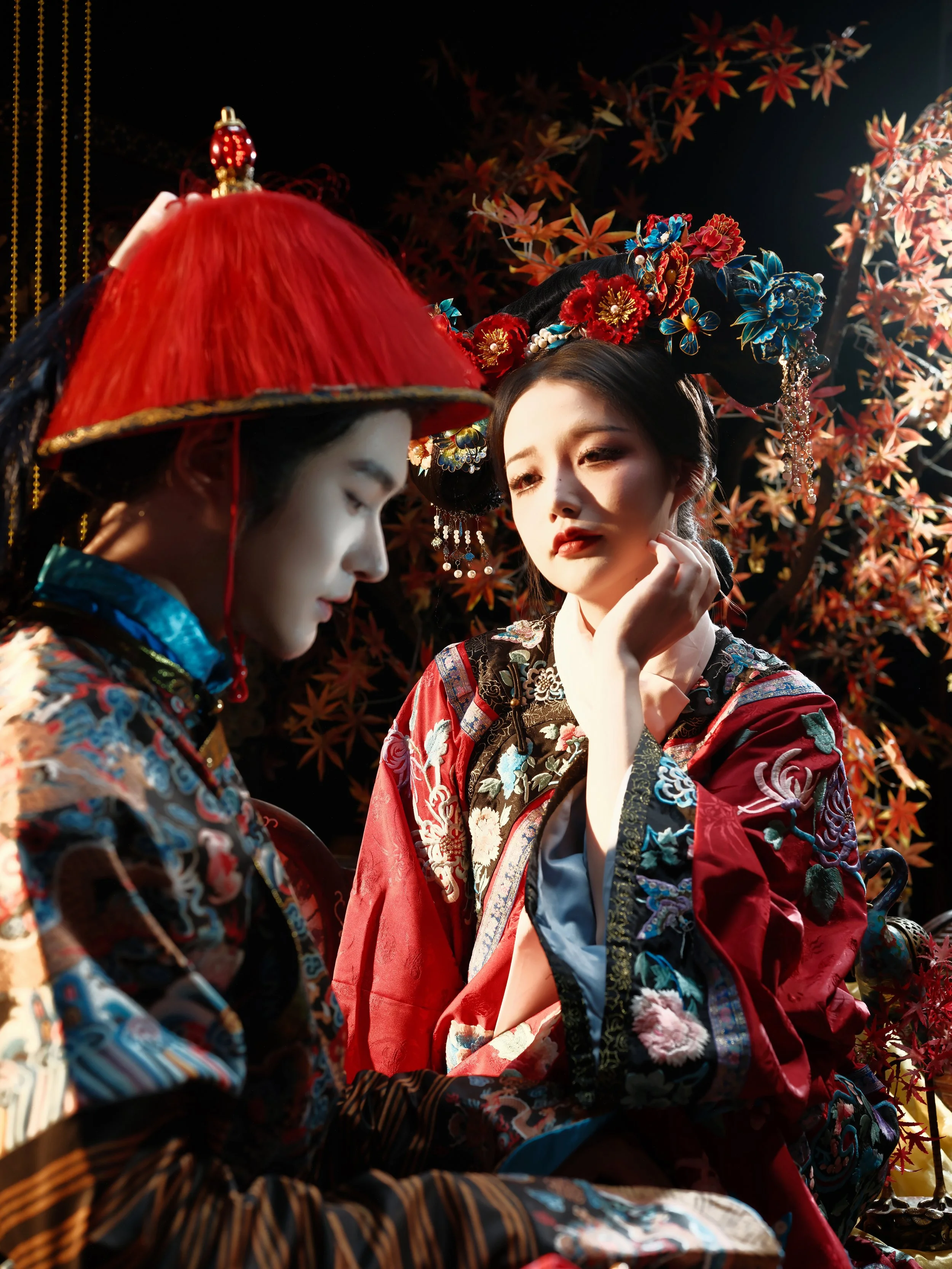 Two women dressed in traditional Japanese kimonos and elaborate hair accessories, with one woman in a red and black kimono wearing a large red hat, and the other in a blue, red, and black kimono with floral embroidery and an ornate black headdress, s