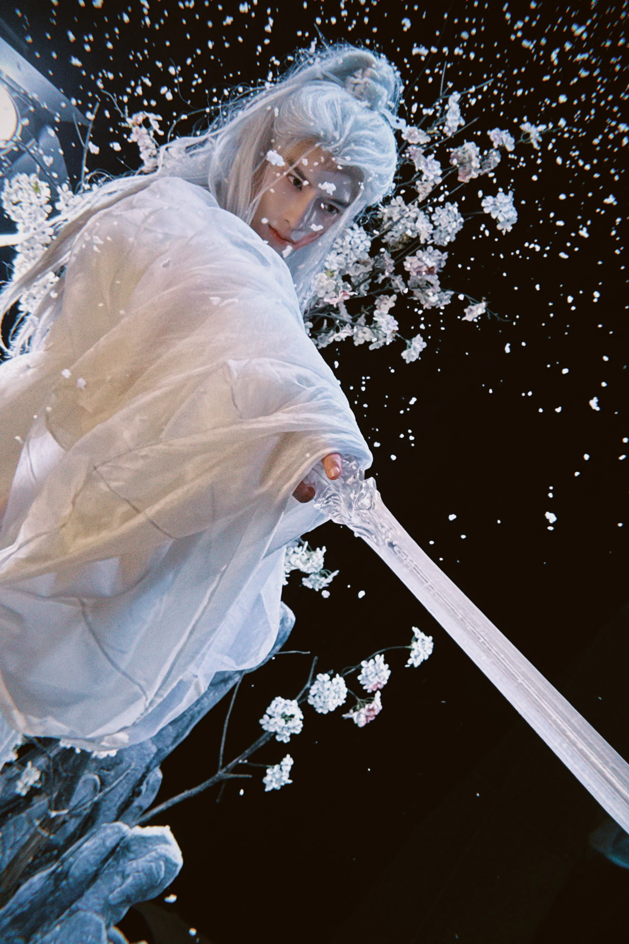 Person dressed in white holding a clear sword, with a background of cherry blossoms and falling petals, against a dark sky.