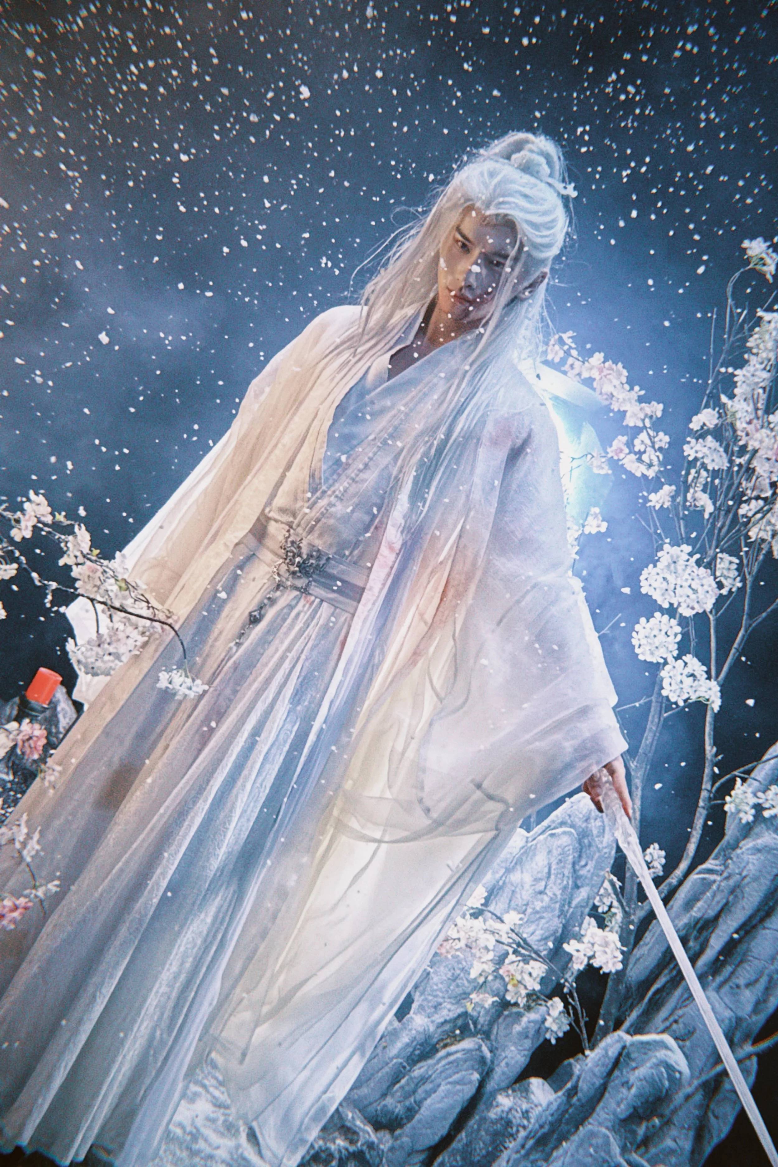 A woman dressed in traditional white robes with long white hair stands outdoors among snow-covered rocks and cherry blossom trees, with snow gently falling around her, illuminated by a bright light behind her.