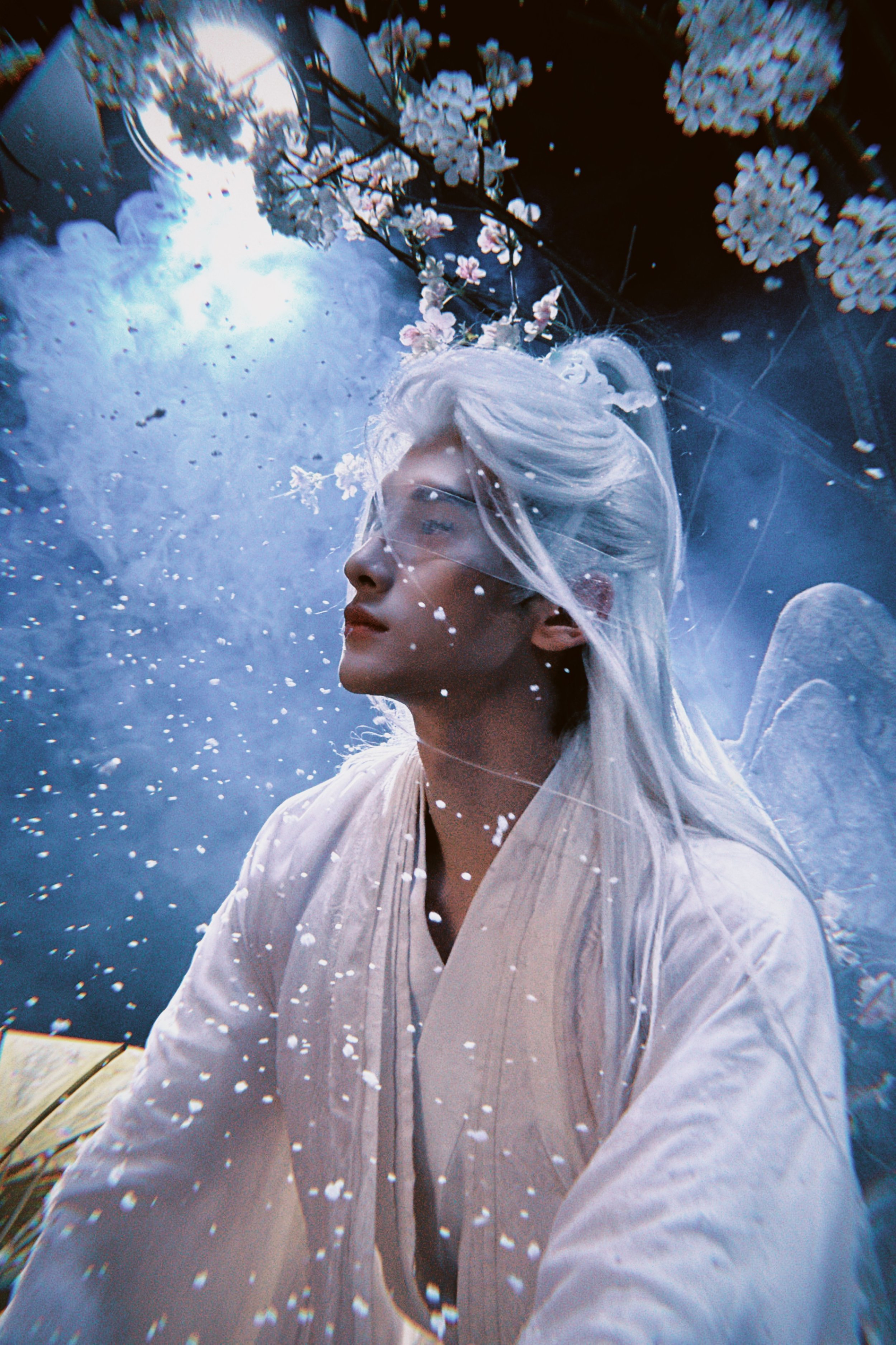 A person with long, white hair and angel wings wearing a traditional cream-colored robe, standing outdoors with cherry blossoms above, surrounded by floating white petals or snow, sunlight filtering through trees.