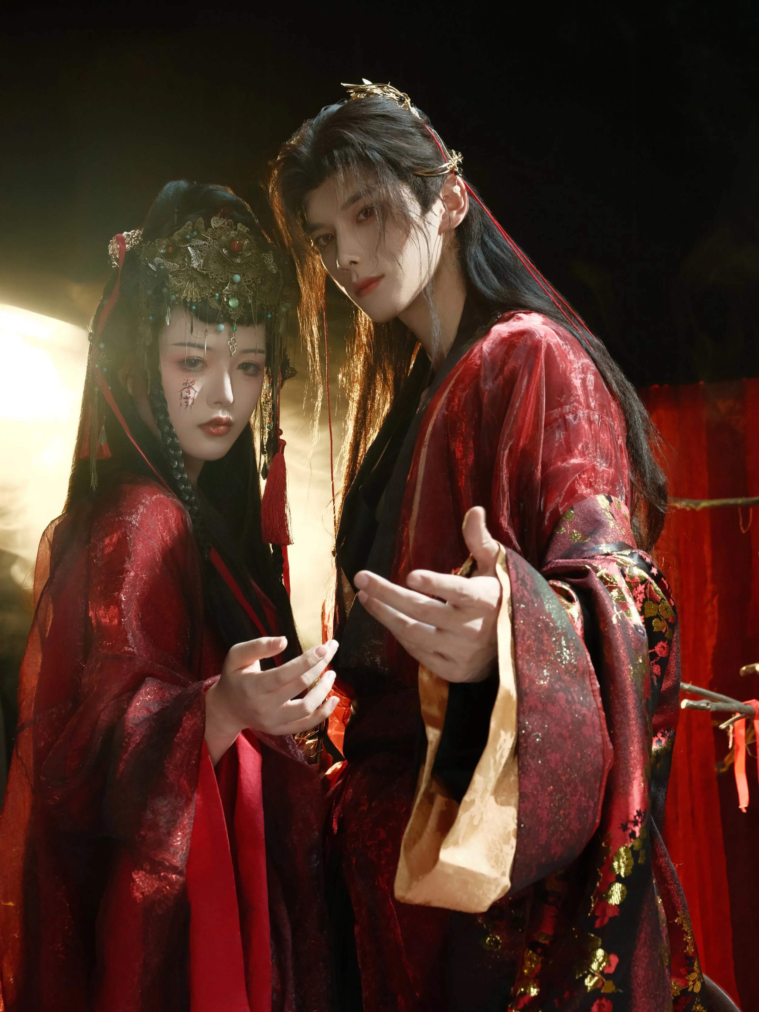 Two individuals dressed in elaborate traditional Asian costumes, with intricate accessories and makeup, standing close together in a dimly lit setting.