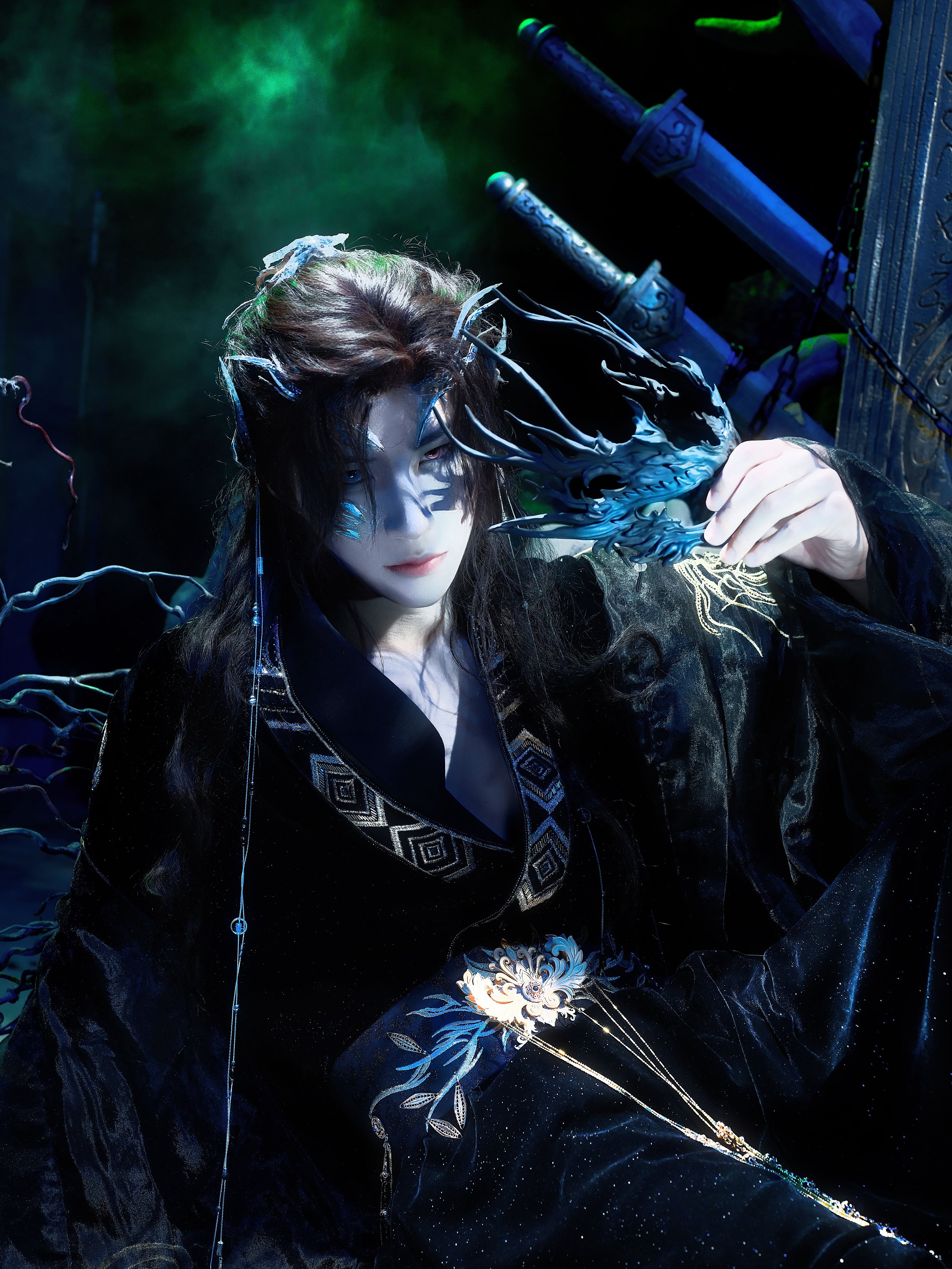 A person dressed in fantasy costume with intricate details, holding a black and blue claw-like prop near their face, surrounded by a dark, mystical background with swords and glowing effects.