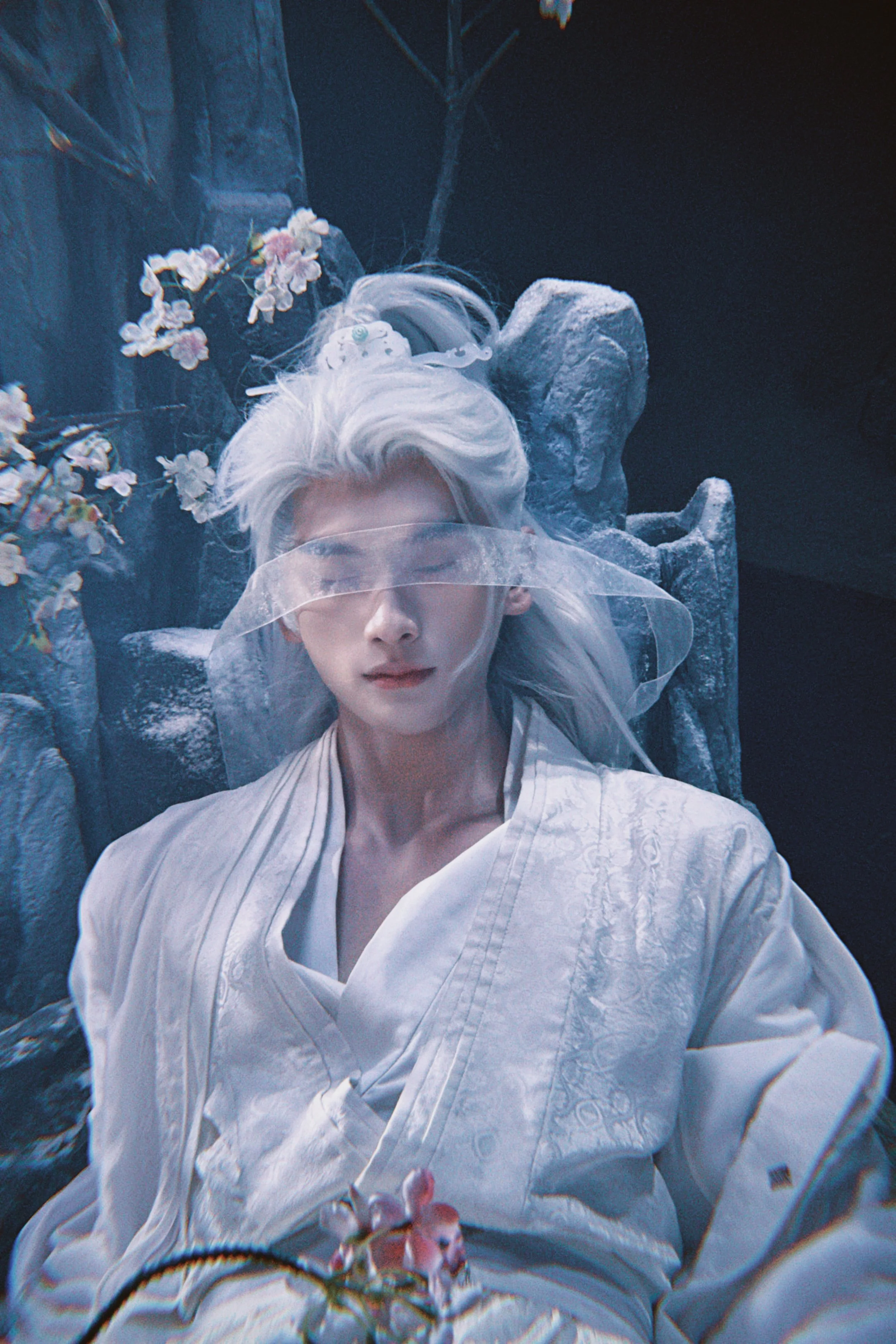 A person with long silver hair lying down with eyes closed, wearing a white embroidered garment and a clear face shield, surrounded by flowers and stone-like structures.
