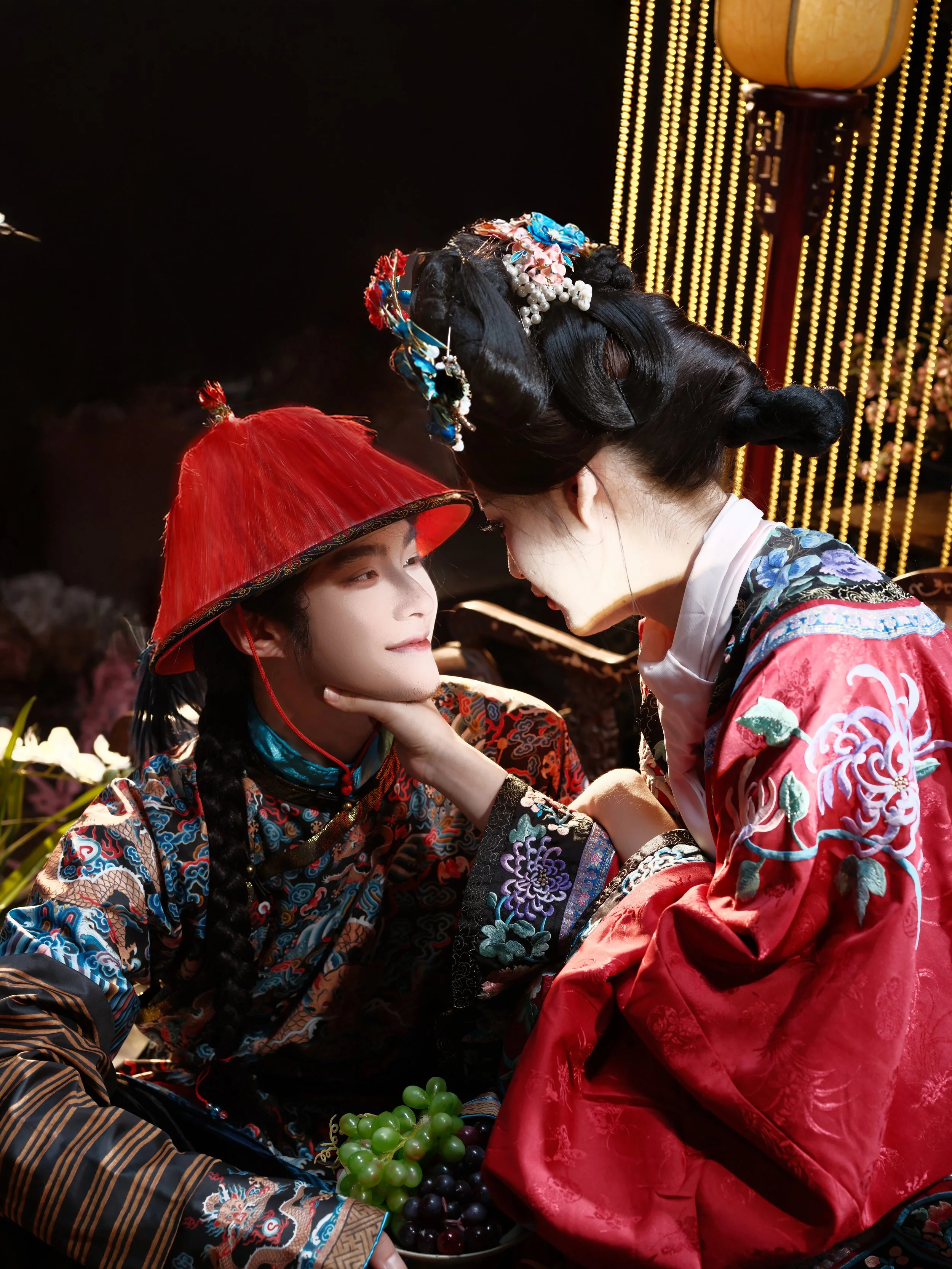 Two people dressed in traditional Asian clothing, one wearing a red conical hat and the other with elaborate hair accessories, are engaged in a close, intimate moment. The person with the conical hat looks at the other with a gentle expression, while