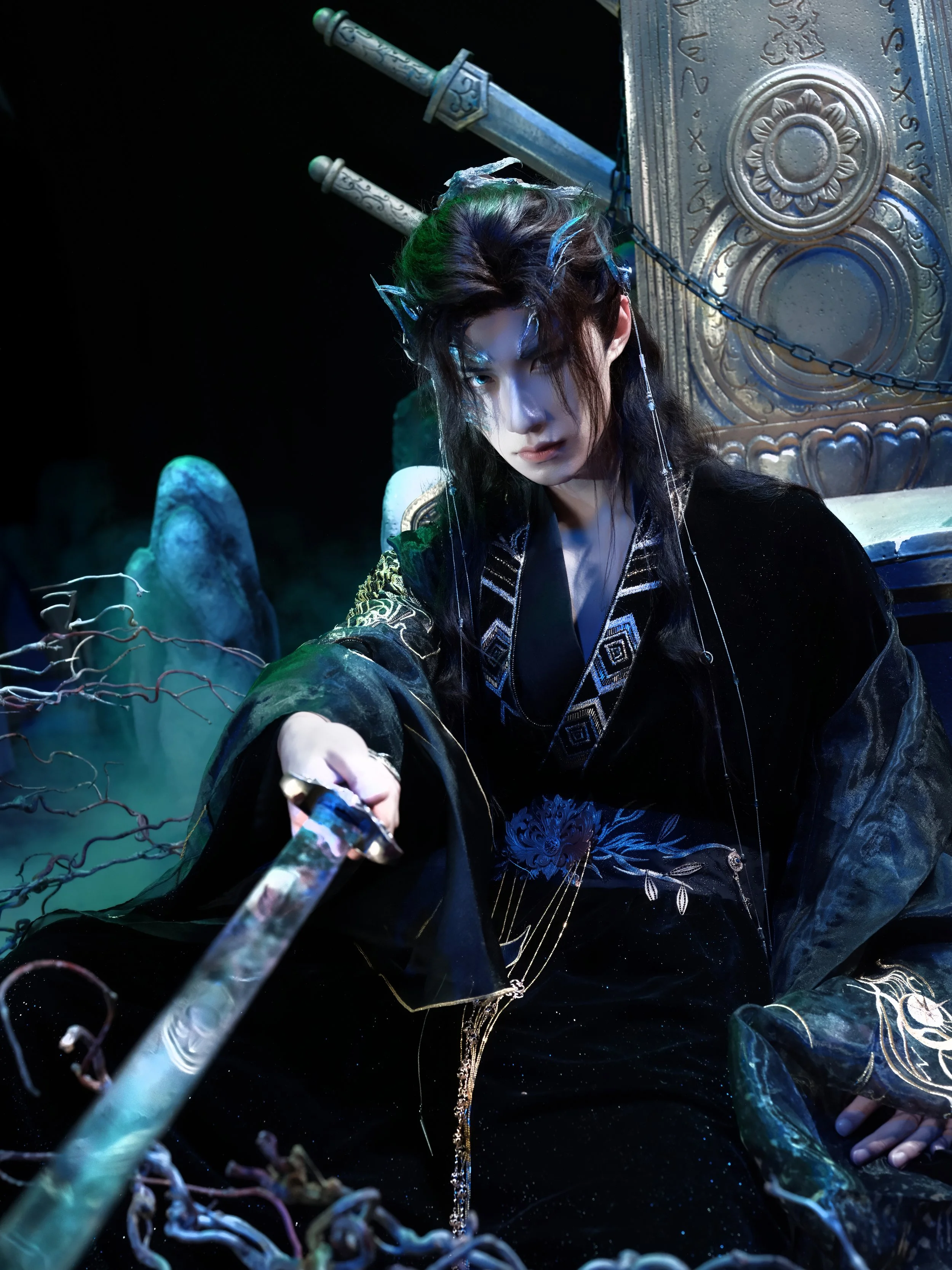 A person dressed in elaborate fantasy costume, sitting on a throne in a dark setting with mystical props and chains.