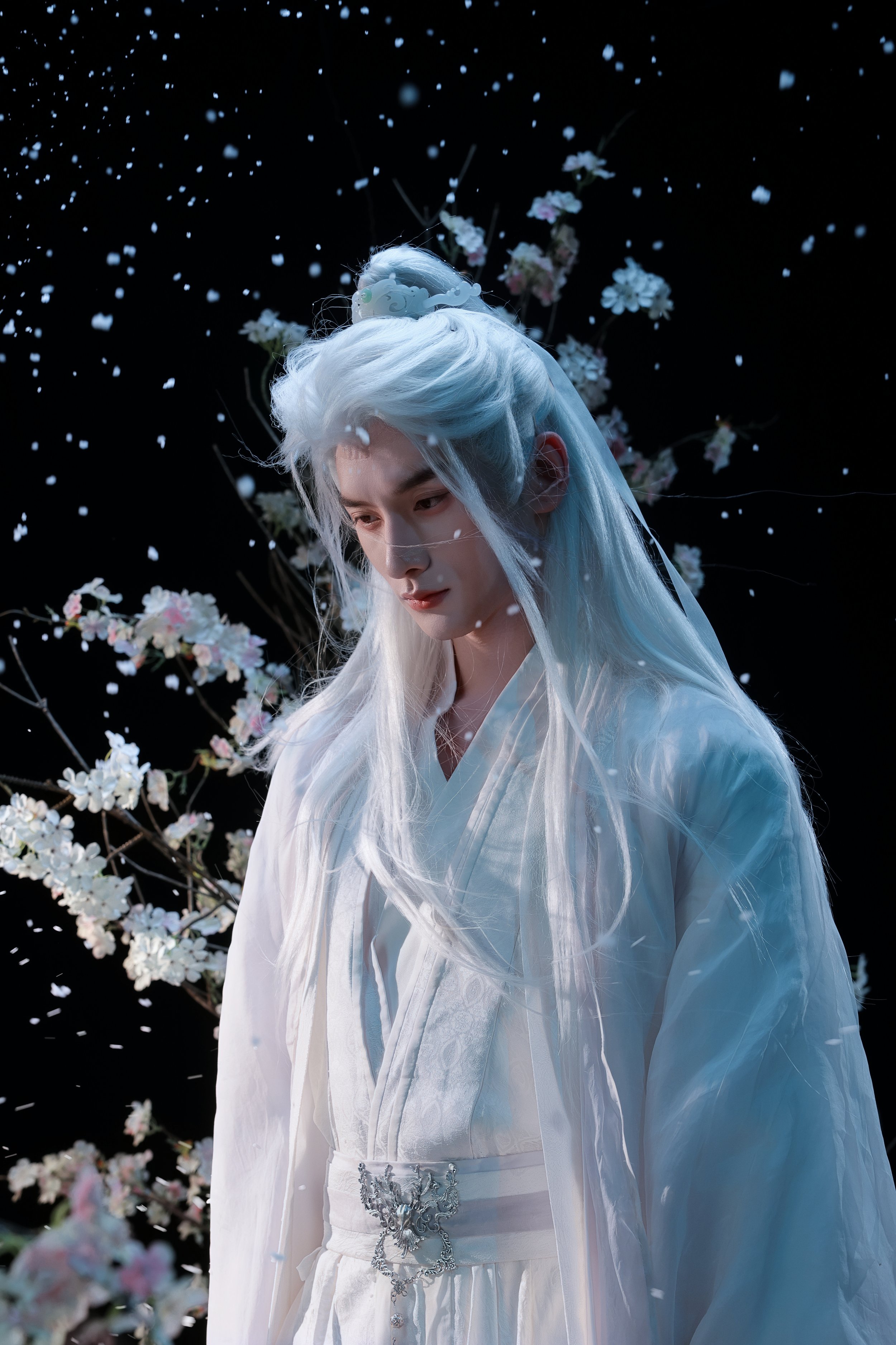 Person dressed in traditional white clothing with long, flowing white hair, standing among cherry blossoms with falling petals against a dark background.