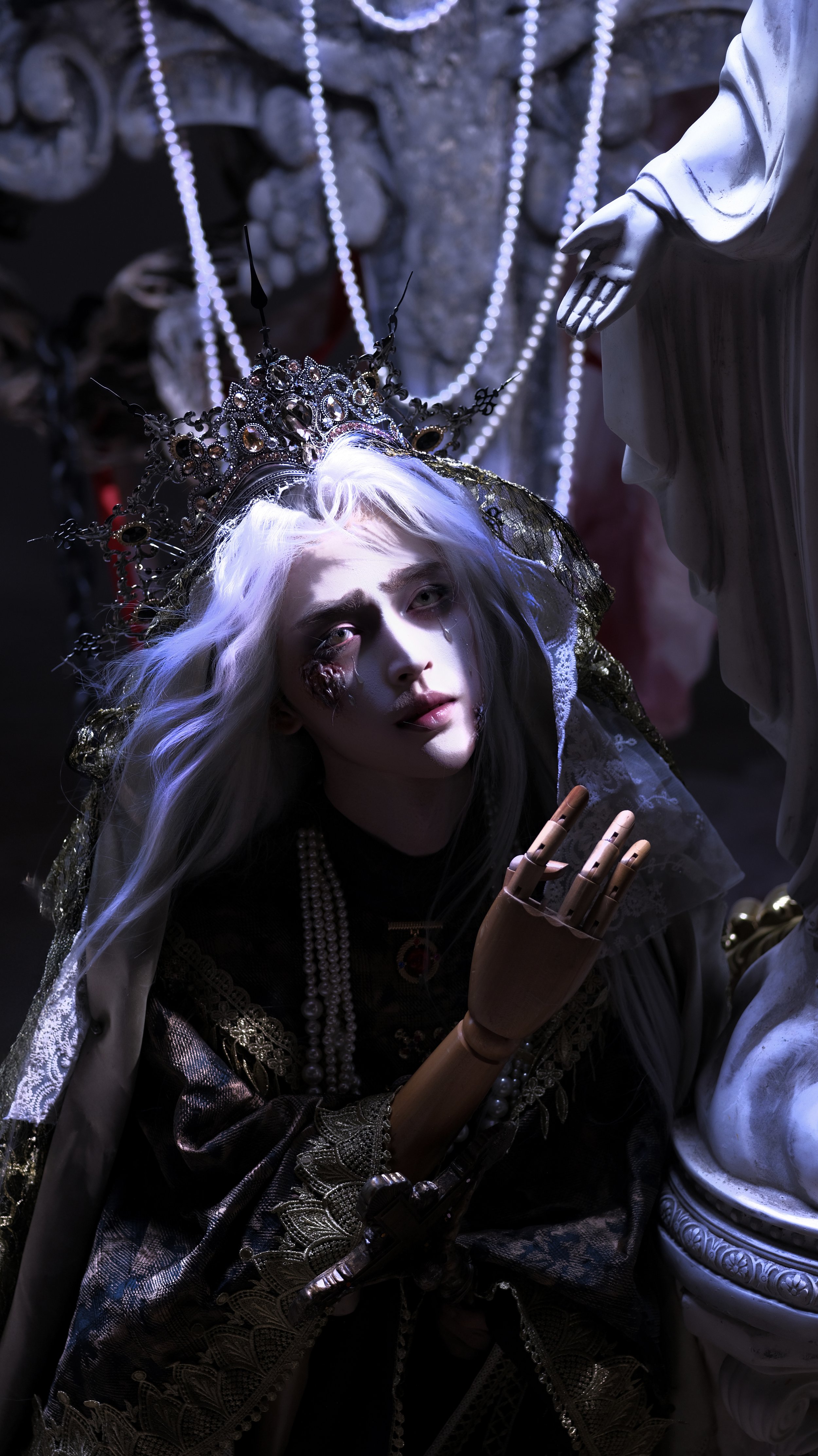 A person dressed as a gothic queen with a crown, white hair, and dramatic makeup, kneeling next to a religious statue in a dimly lit setting.