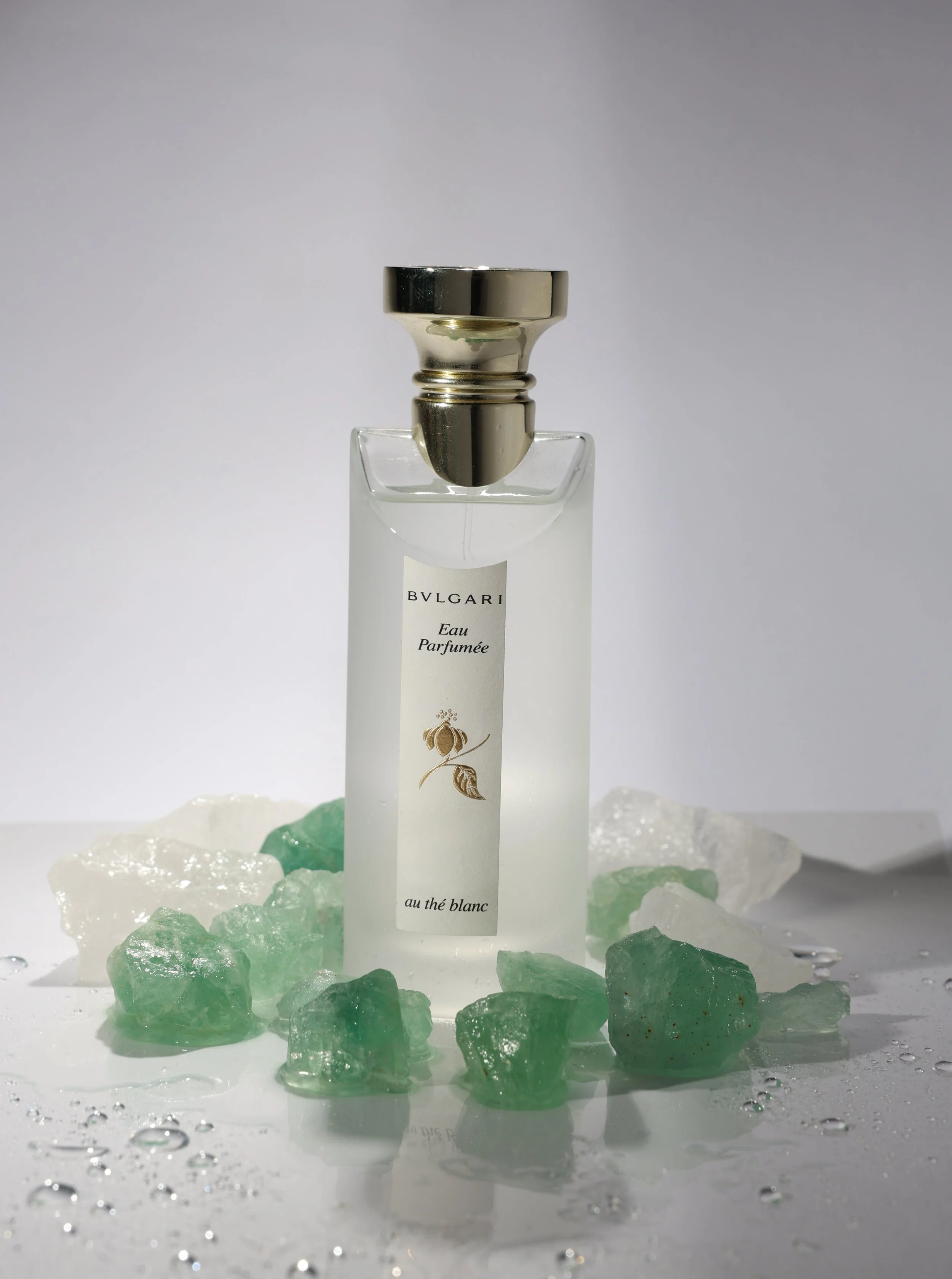 A bottle of BVLGARI Eau Parfumée perfume surrounded by green and white stones and some water droplets.