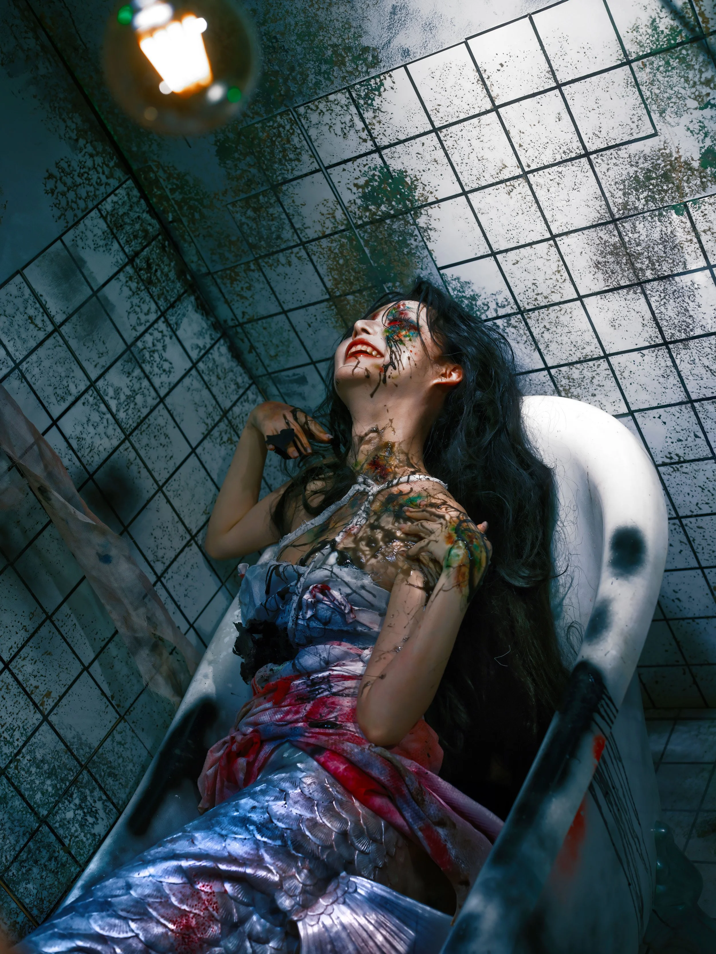 A woman with long black hair and colorful face paint, dressed as a mermaid with a tail, lying in a stained bathtub in a tiled bathroom with a dirty mirror.