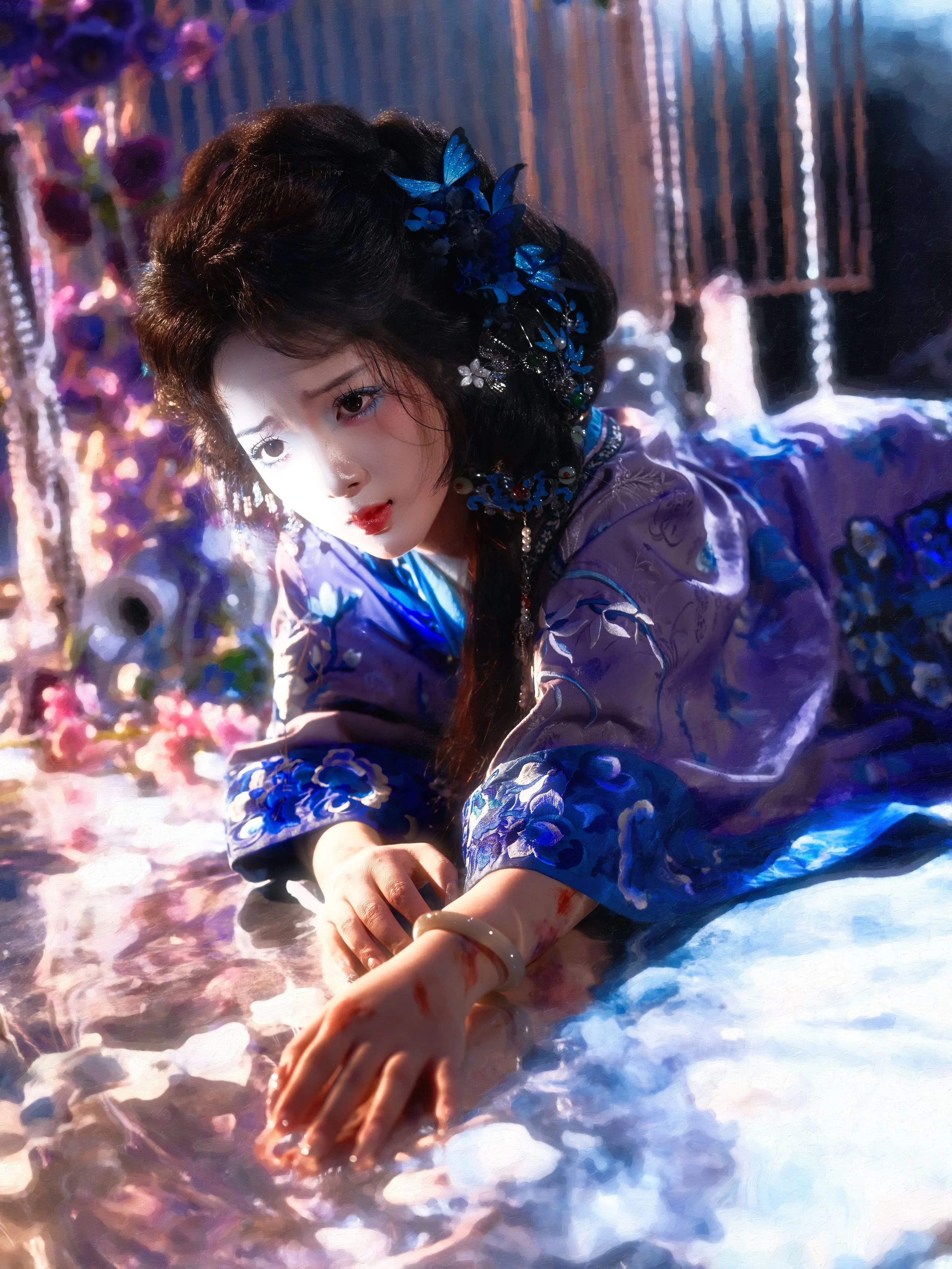 A woman dressed in a purple traditional Asian garment with intricate blue embroidery, lying on the floor with her hand extended. She has dark hair decorated with blue flowers and jewelry, with a background of brightly colored flowers and decorative b