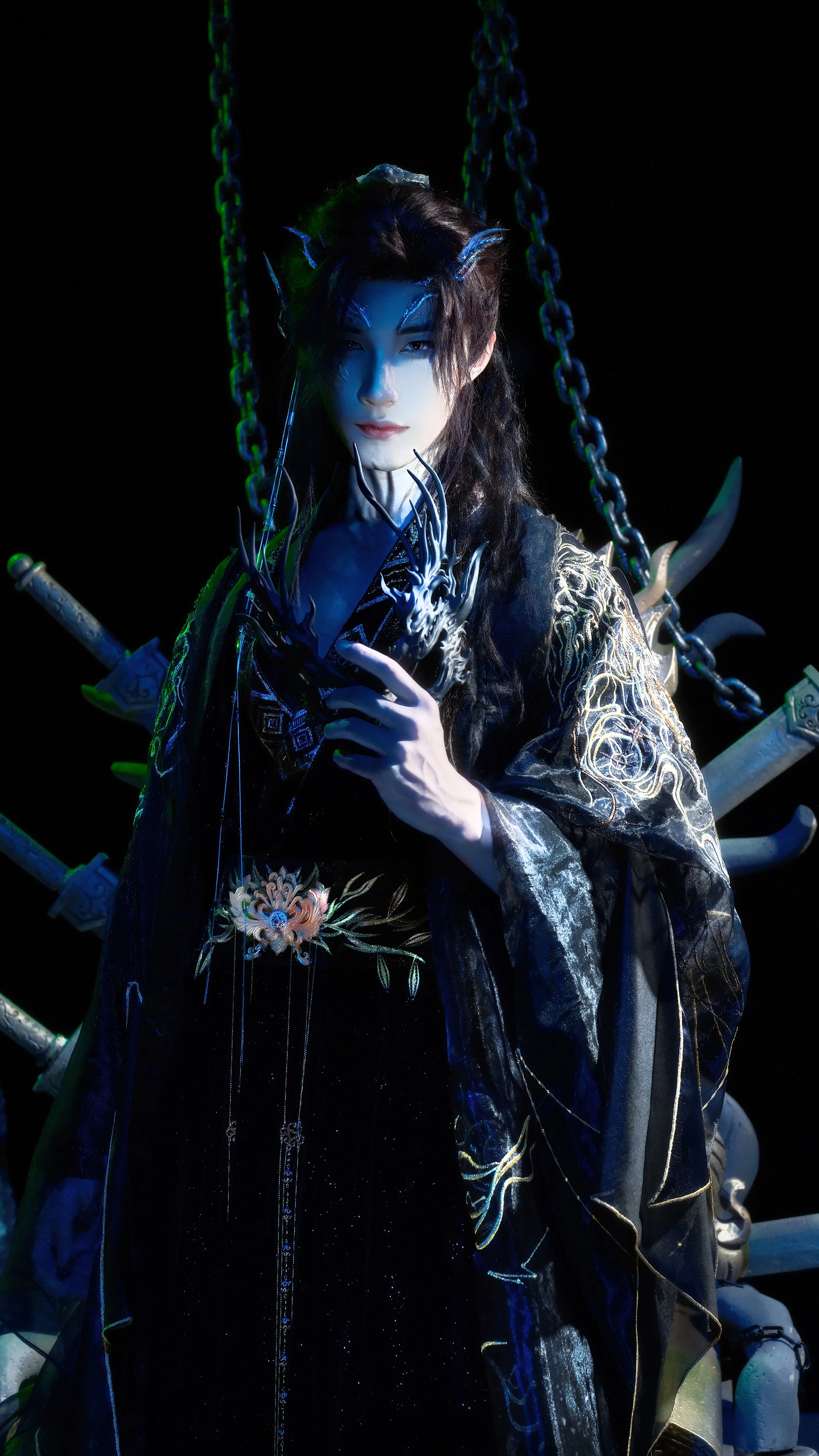 A person dressed in a dark, elaborate costume with gold embroidery, holding a decorative mask, standing in front of a backdrop with chains and swords, illuminated by dramatic lighting.