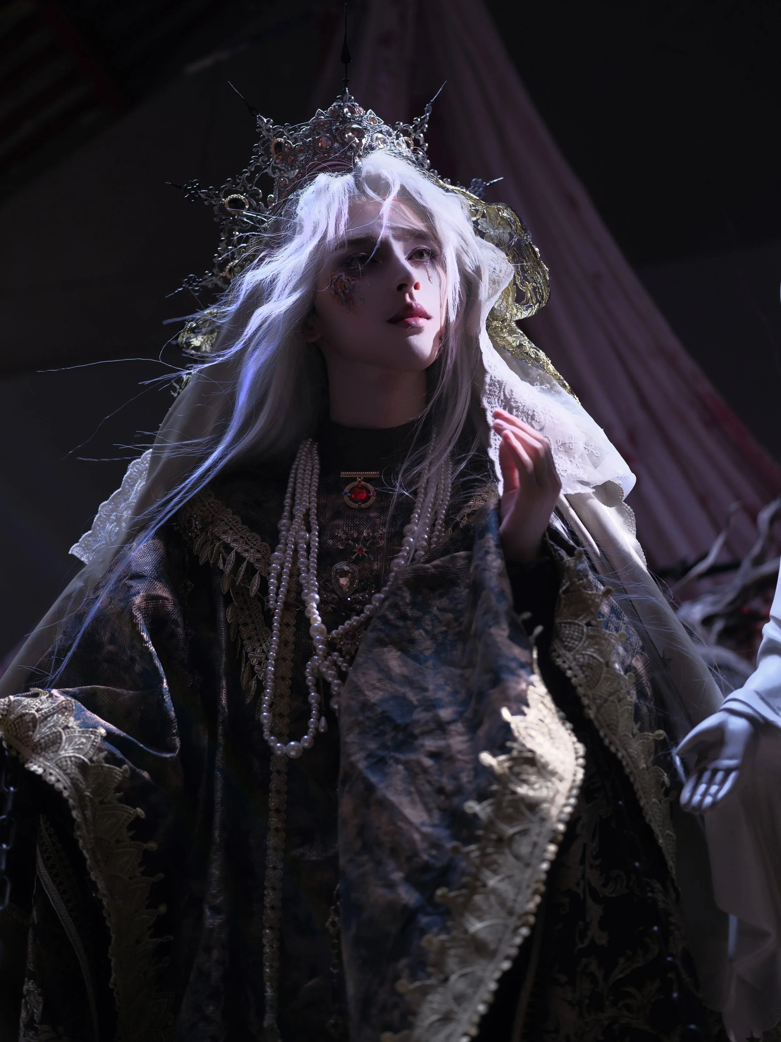 A woman dressed in elaborate, dark, regal clothing with layered jewelry, including pearls and a red gem necklace, wearing a silver crown and a veil, with long white hair, in a dimly lit setting.