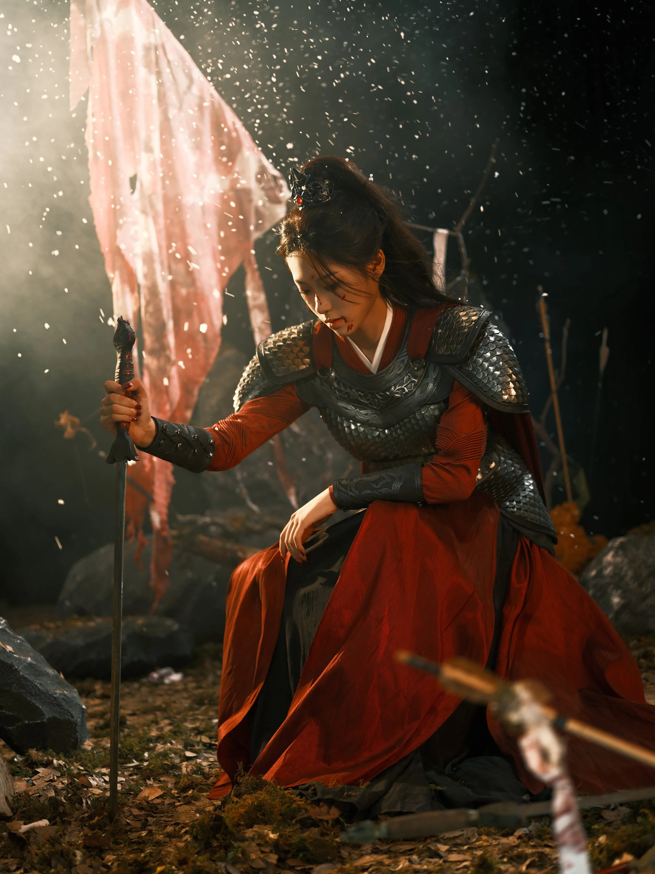 A woman dressed in medieval armor and a red skirt kneels on a forest floor, holding a sword with her eyes closed, surrounded by falling leaves and a flag in the background.