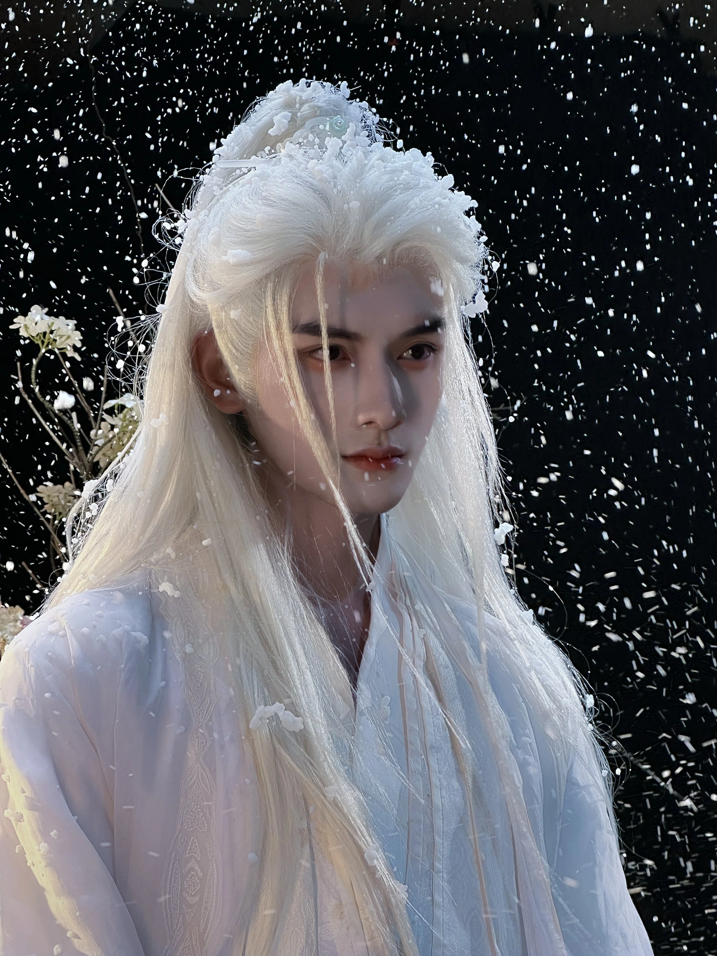 A person with long, platinum blonde hair and fair skin stands in the snow at night, wearing a white garment, with snow falling around them and a dark background.