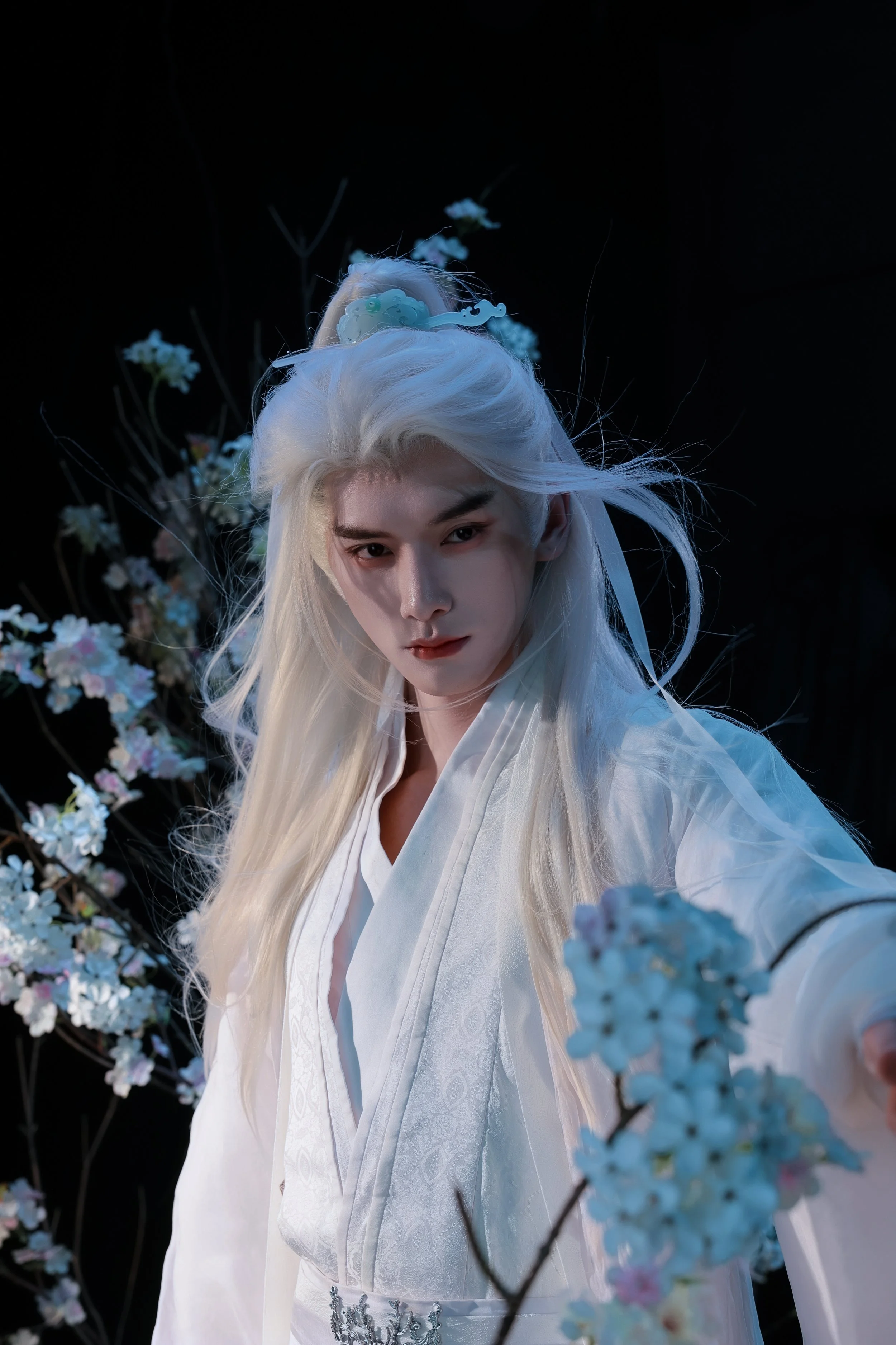 A person dressed in white with long white hair standing among pastel-colored flowers in a dark setting.