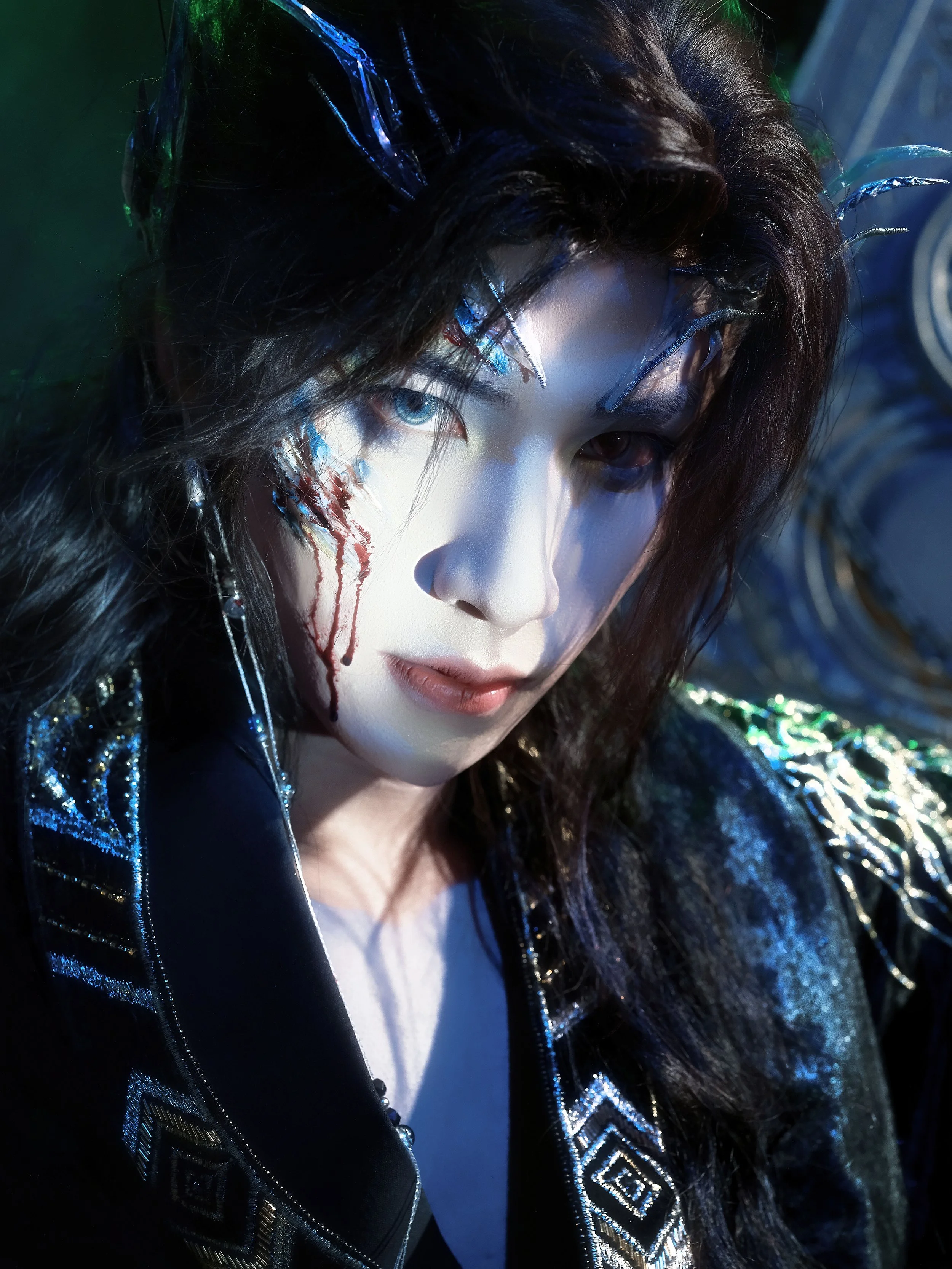 A person with dark, wavy hair and striking makeup, including metallic blue and silver accents around the eyes and red streaks on the face, wearing a shiny black jacket with intricate patterns.