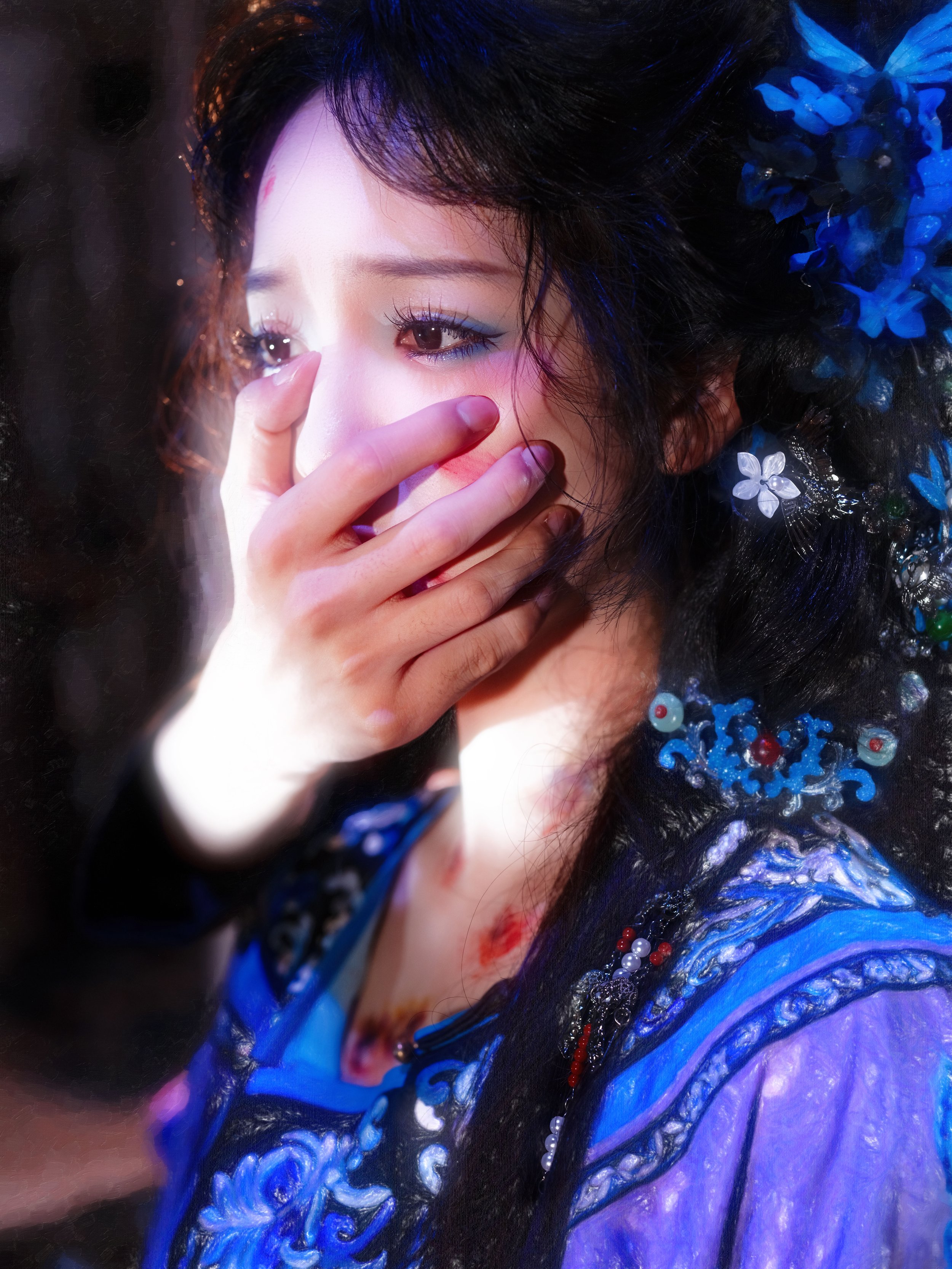 A woman with dark hair and traditional makeup, wearing a blue embroidered outfit, covering her mouth with her hand, with floral hair accessories.