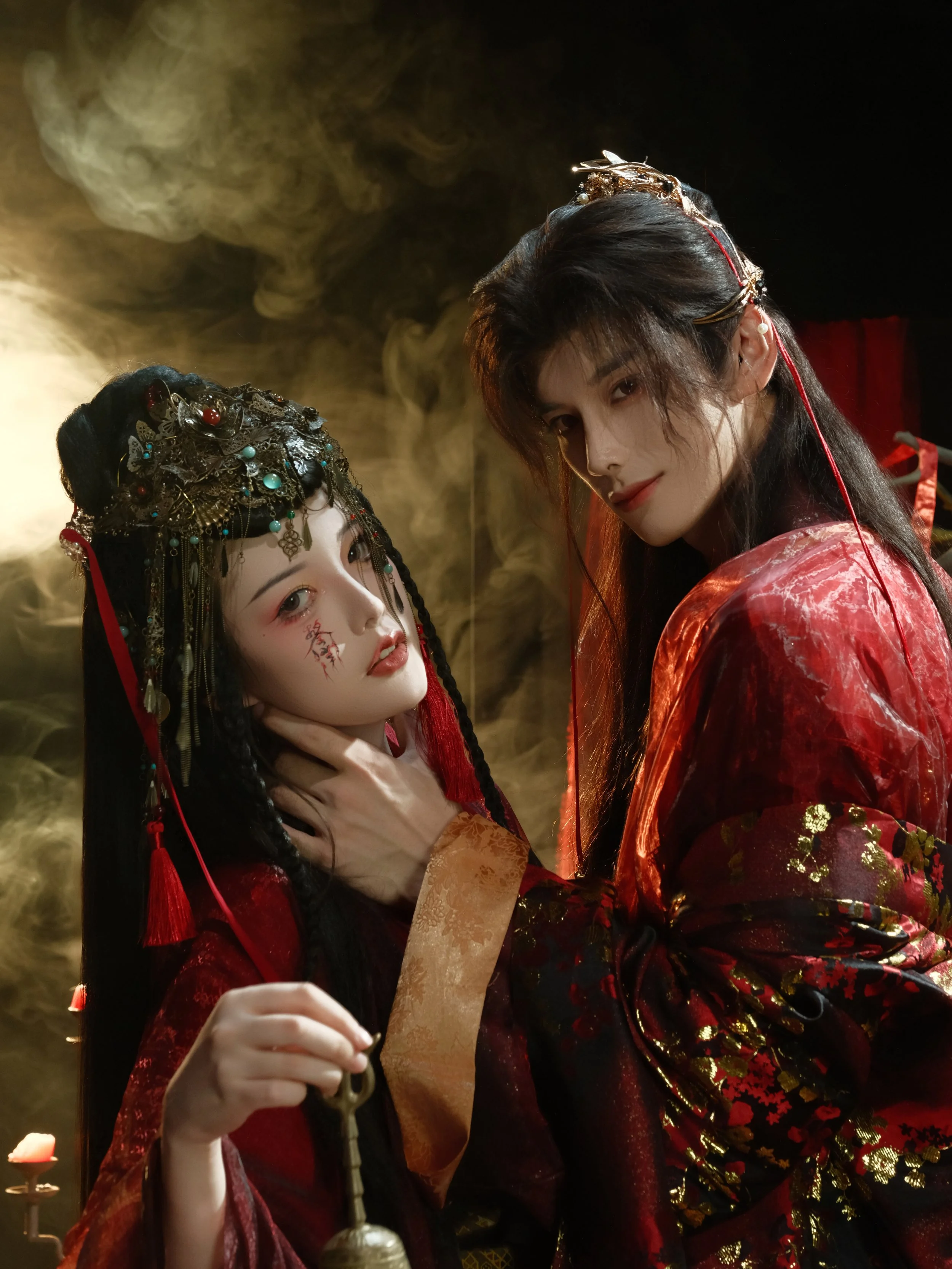 Two people in elaborate, traditional Asian-style costumes with intricate jewelry and headpieces, standing against a smoky background, creating a dramatic, mysterious atmosphere.
