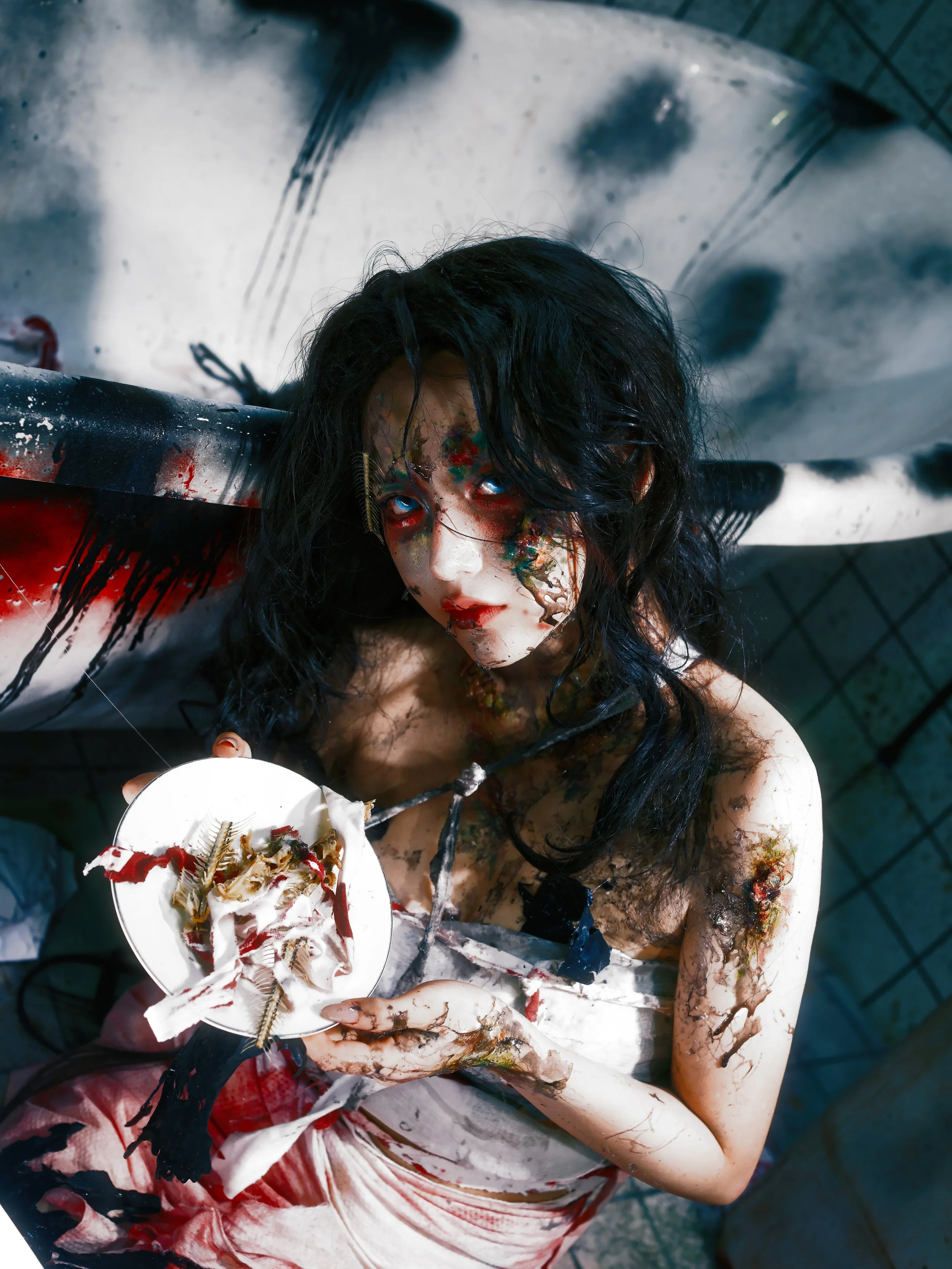 A woman with messy black hair, smeared makeup, and a blood-stained dress, lying on a tiled floor with a large, stained, white pipe behind her, holding a torn plate of food with insects.