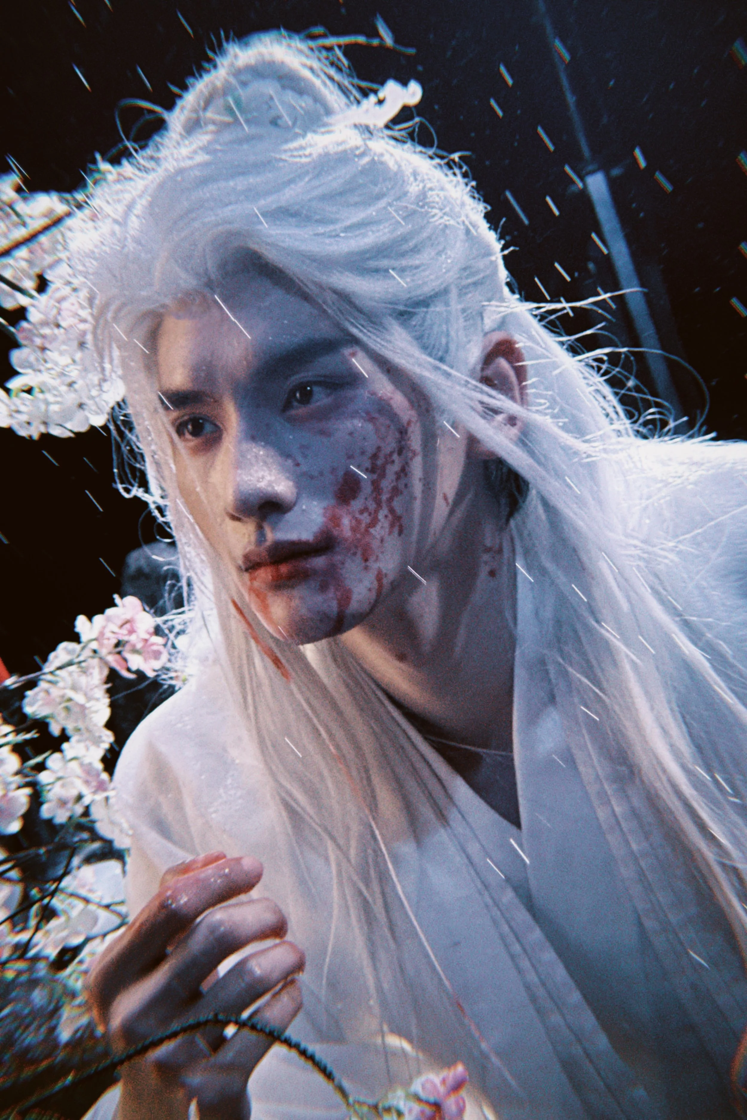A person with long platinum hair and makeup resembling blood on their face, wearing white traditional clothing, surrounded by white and pink flowers, in a dark setting with rain or streaks of light.