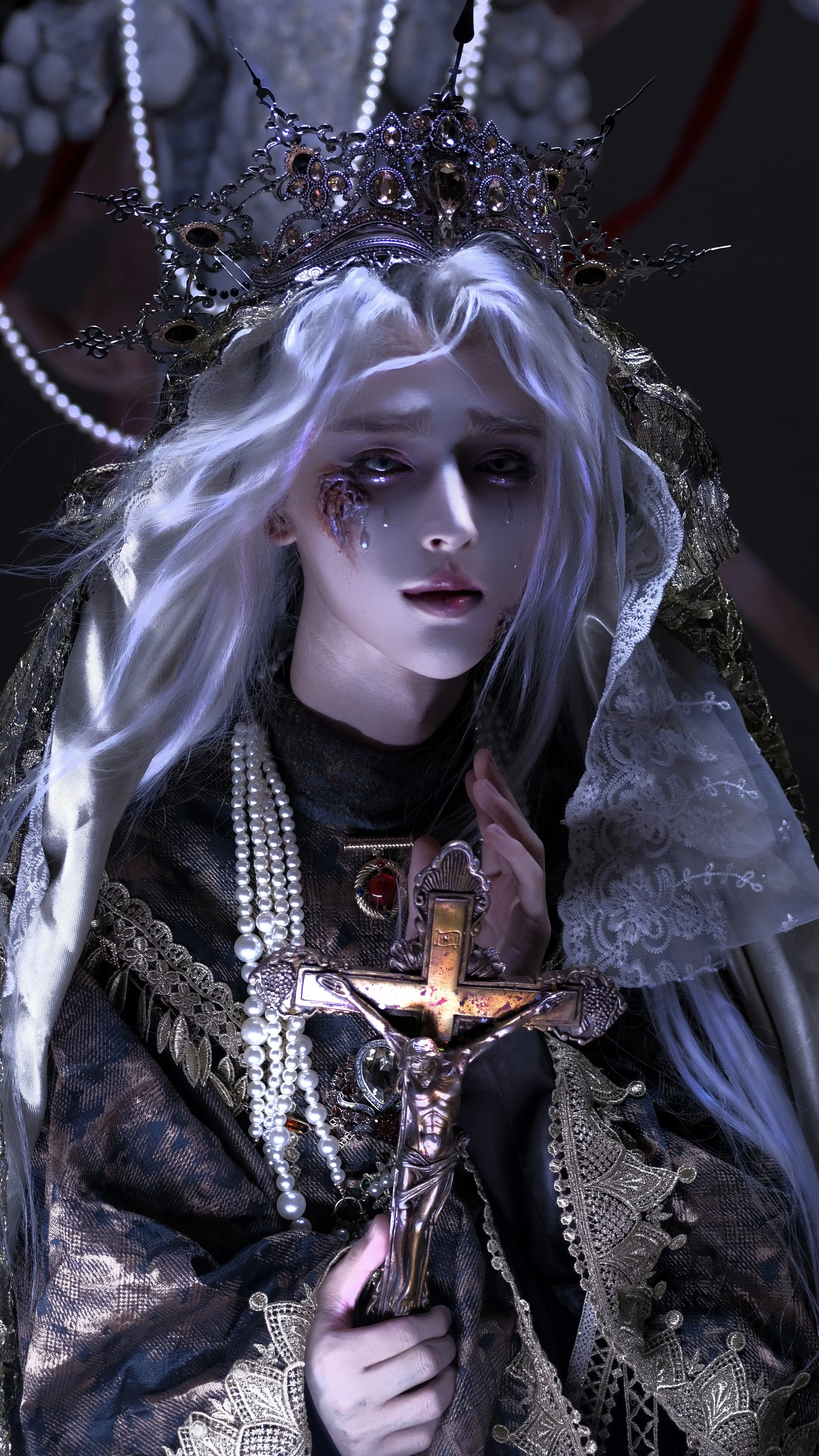 A person with supernatural features, long white hair, wearing a crown, holding a crucifix, with jewelry, lace, and makeup that looks gothic or religious, with dark makeup and tears.