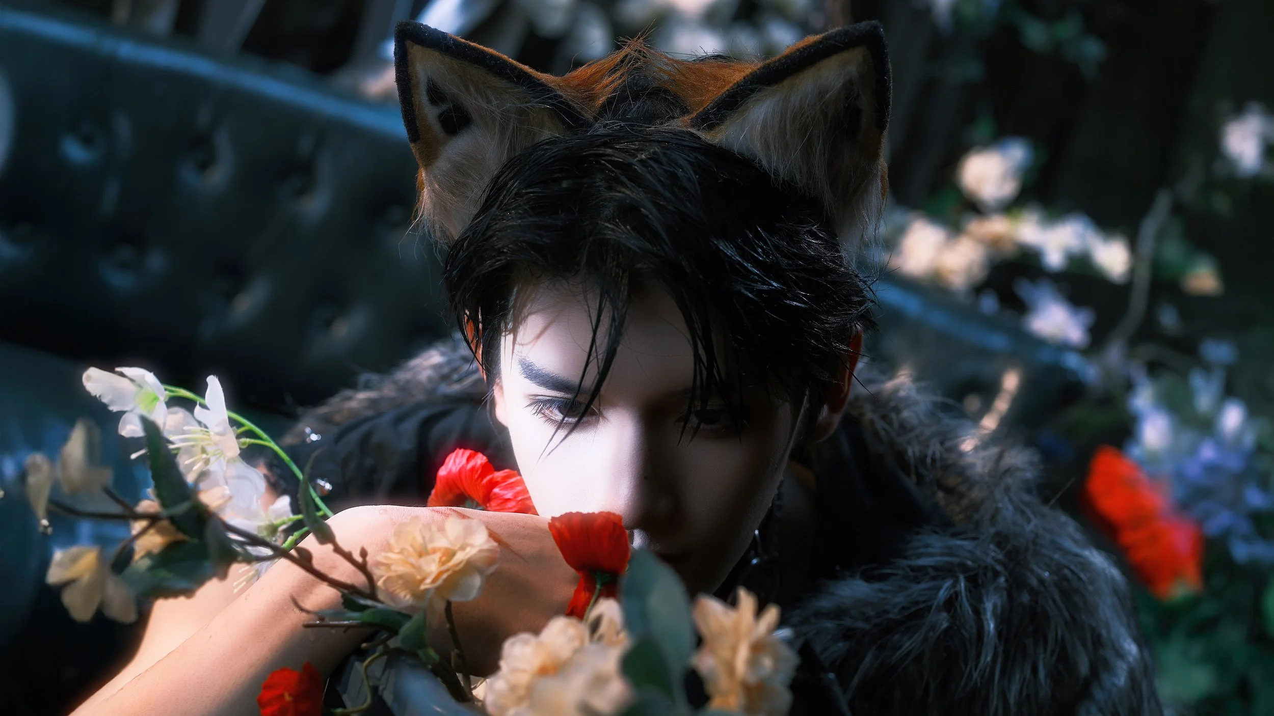 A person with black hair, wearing a furry coat, is surrounded by flowers and has a fox mask on their head. They are holding a red flower close to their face, in a dark and lush outdoor setting.