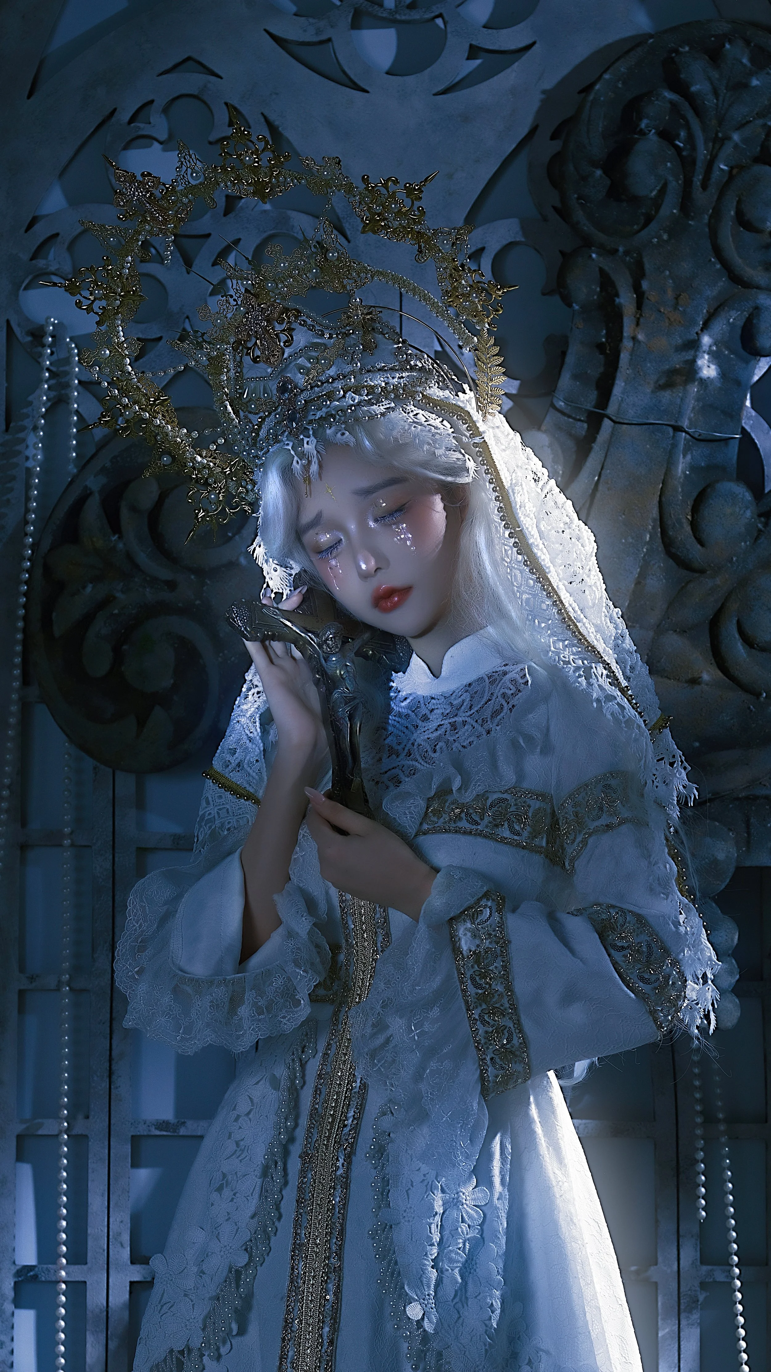 A woman with silver hair, dressed in a white lace gown, holds a crucifix and wears an elaborate gold crown with intricate details, standing against a dark ornately decorated background.
