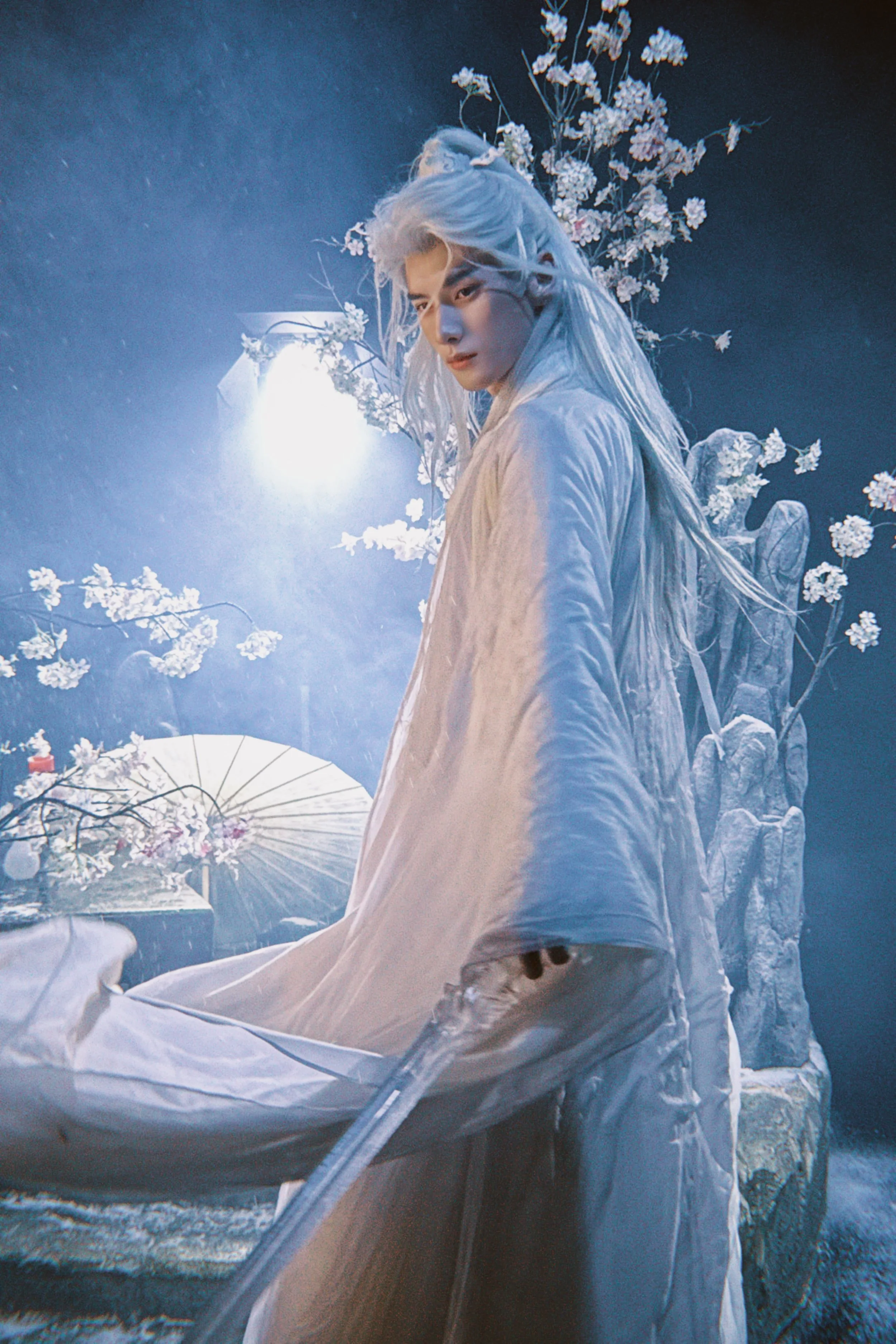 Person in traditional white attire with long white hair, standing in front of a blue background with cherry blossom branches, illuminated by soft lighting.