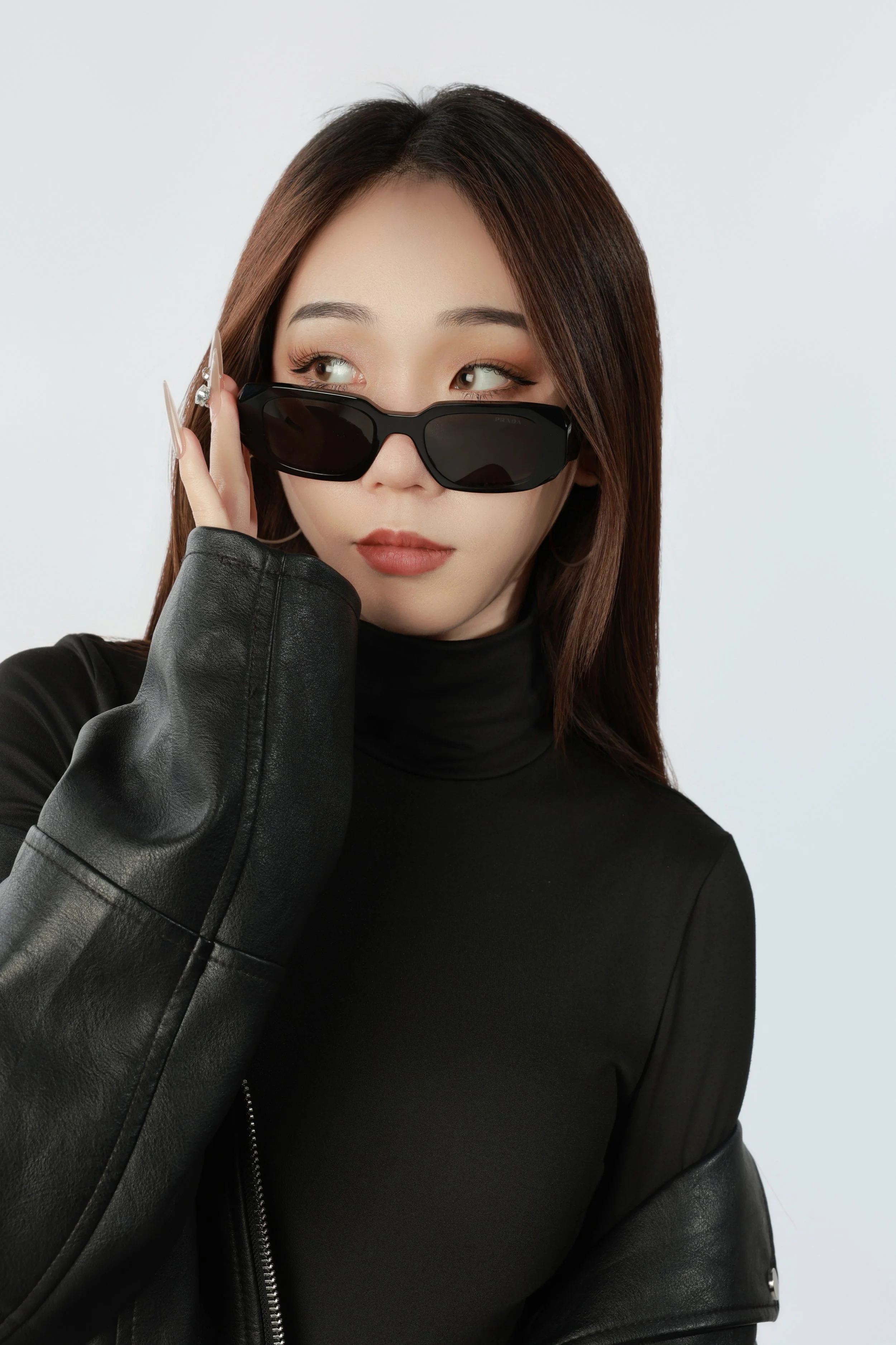 A woman with brown hair wears black sunglasses, a black turtleneck, and a black leather jacket, looking to her left.