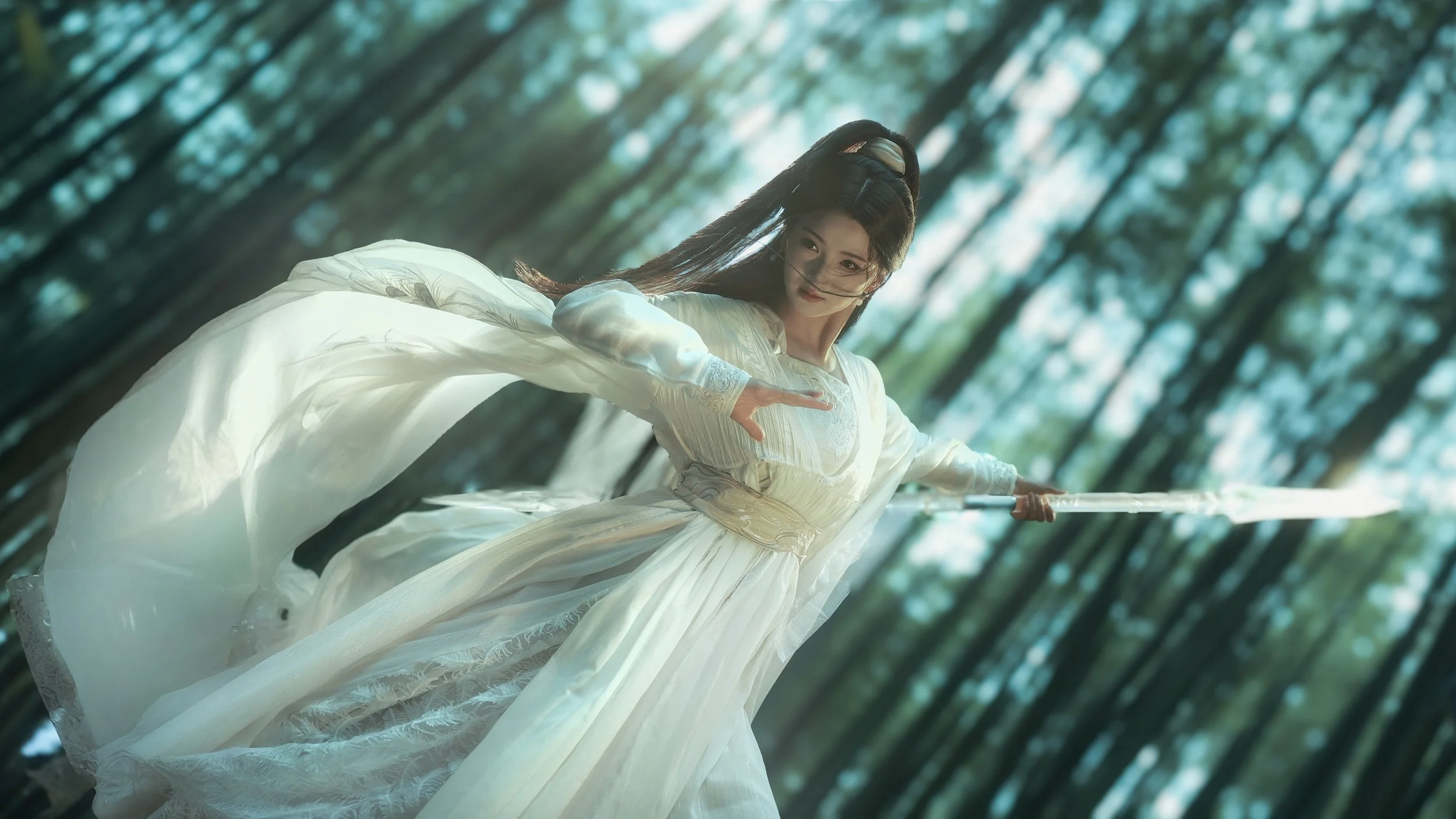 A woman dressed in traditional white flowing attire, holding a sword, stands in a lush green forest with tall trees and sunlight filtering through the leaves.