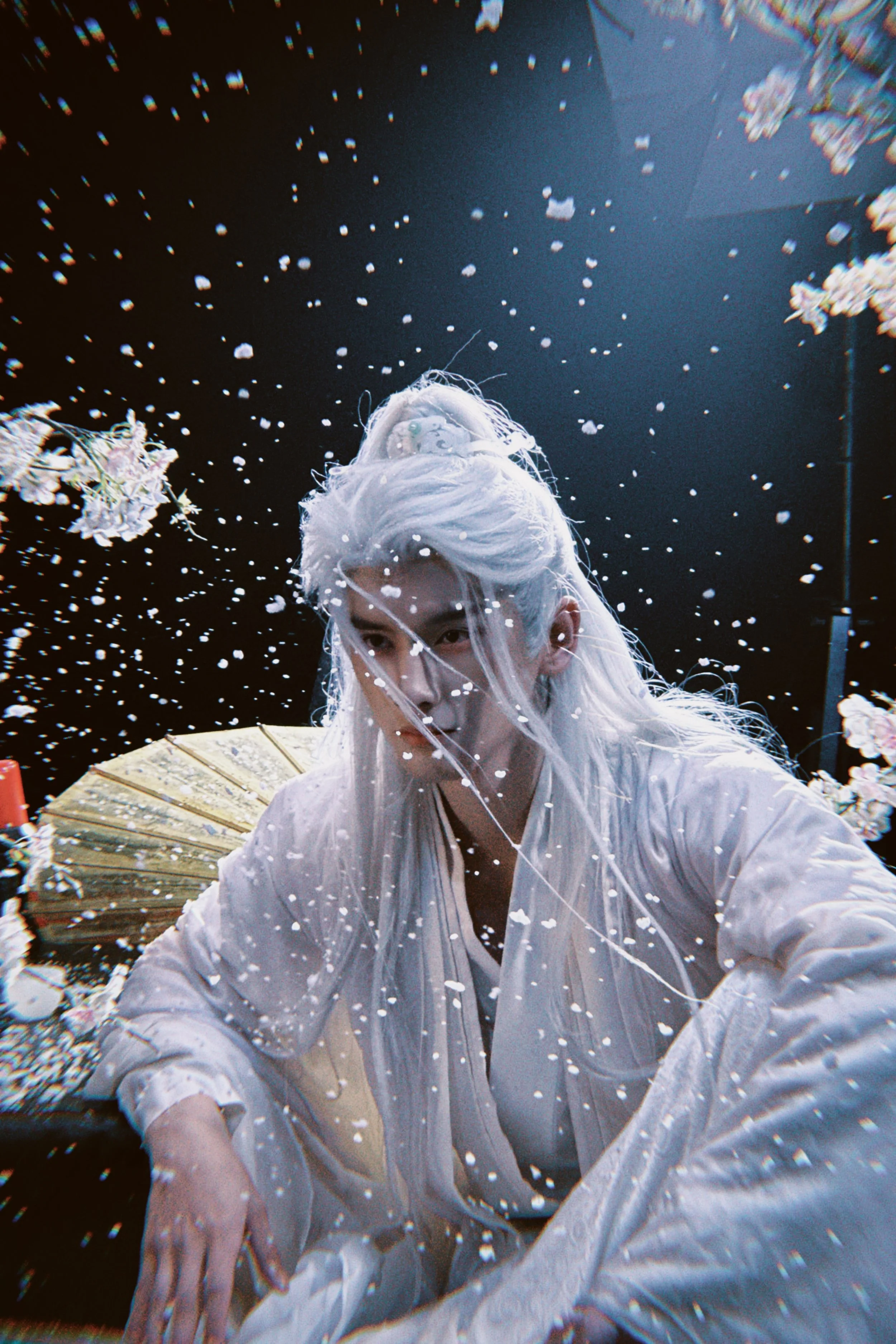 A person with long white hair dressed in white traditional clothing, surrounded by falling cherry blossoms and confetti, holding a closed paper fan, with a dark background.