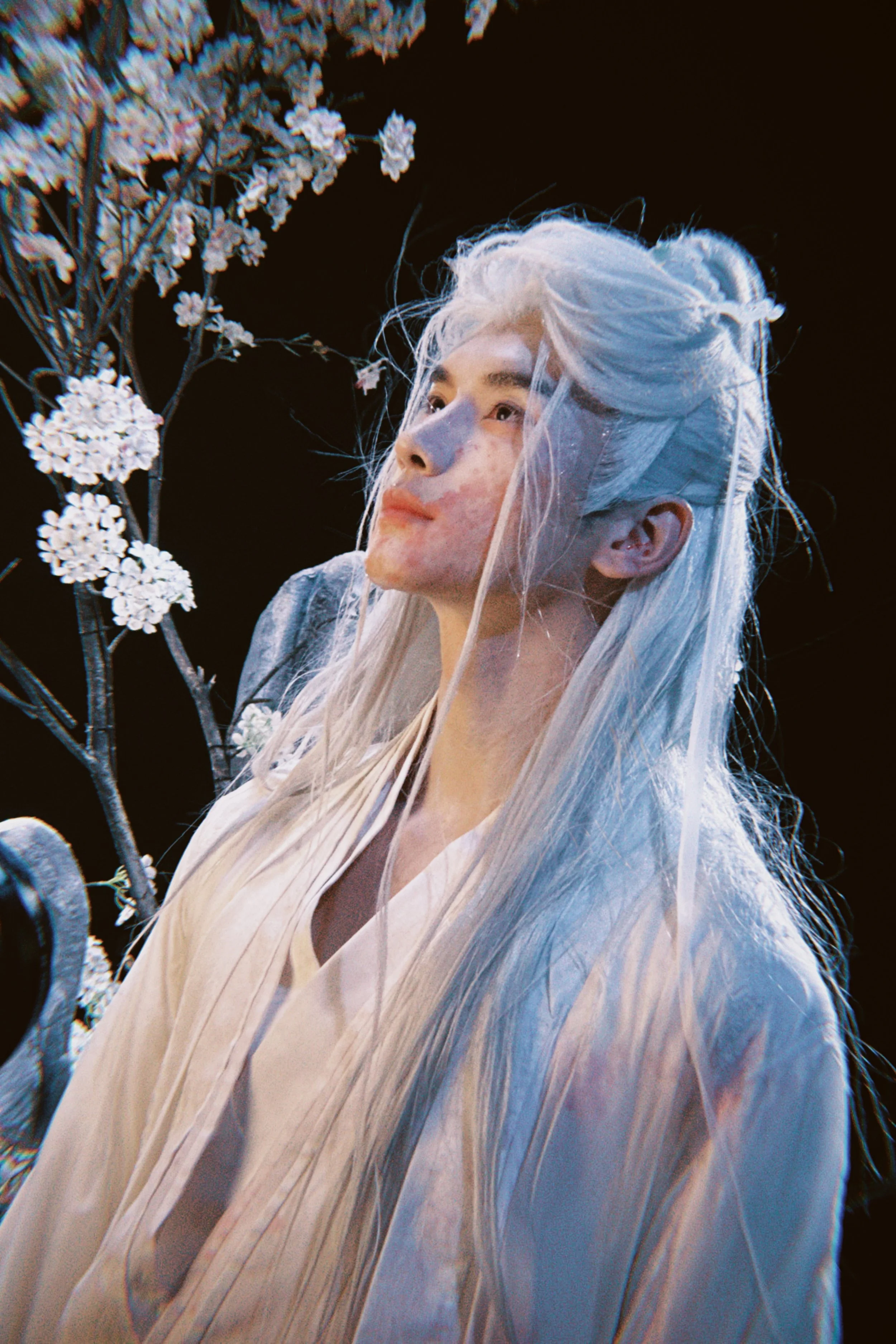 A person with long, pale blue hair and makeup with freckles, standing near a branch with white flowers, against a dark background.