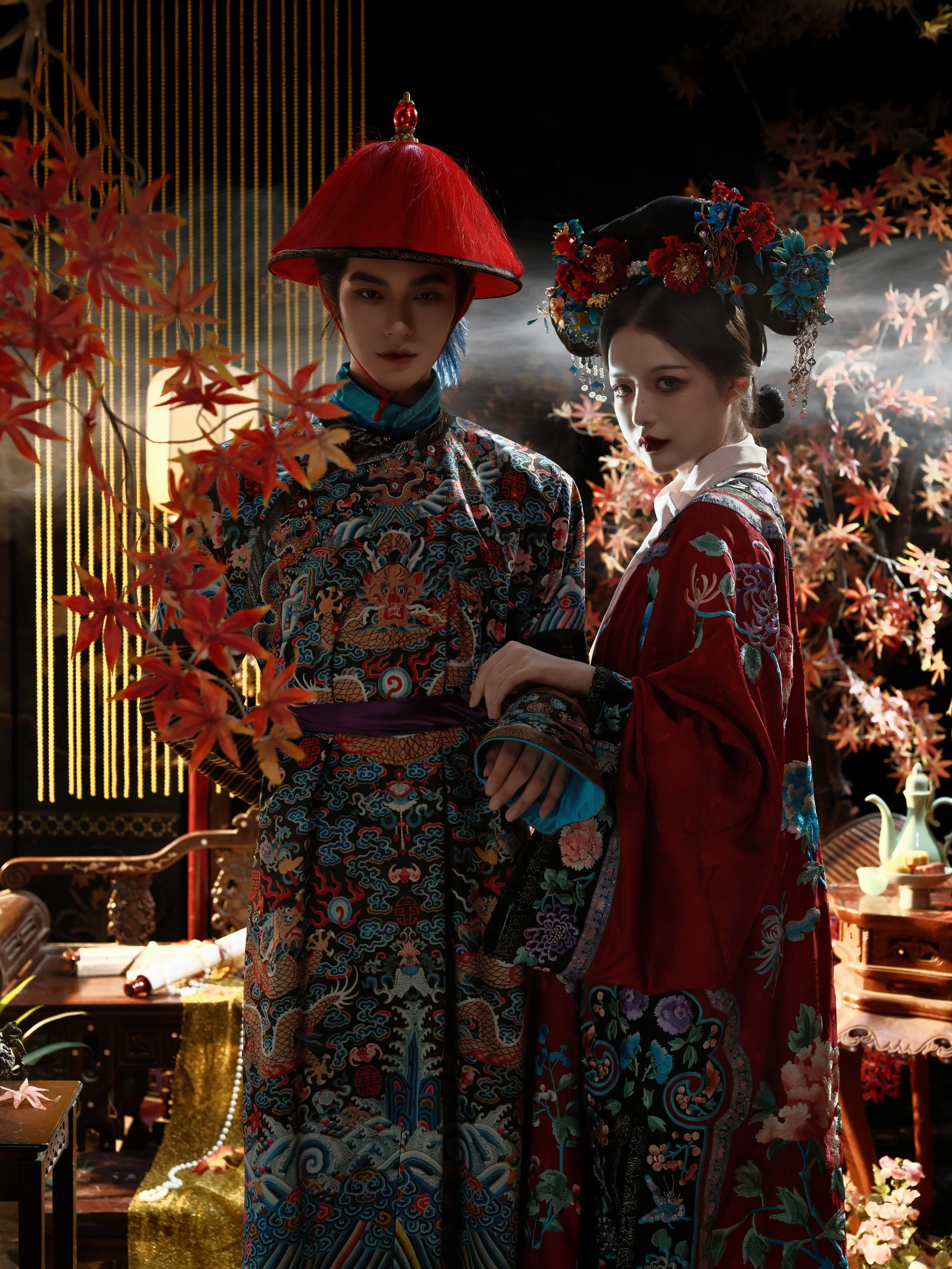Two women dressed in traditional Asian garments with intricate embroidery, standing amidst a decorated setting with autumn leaves and traditional furniture.