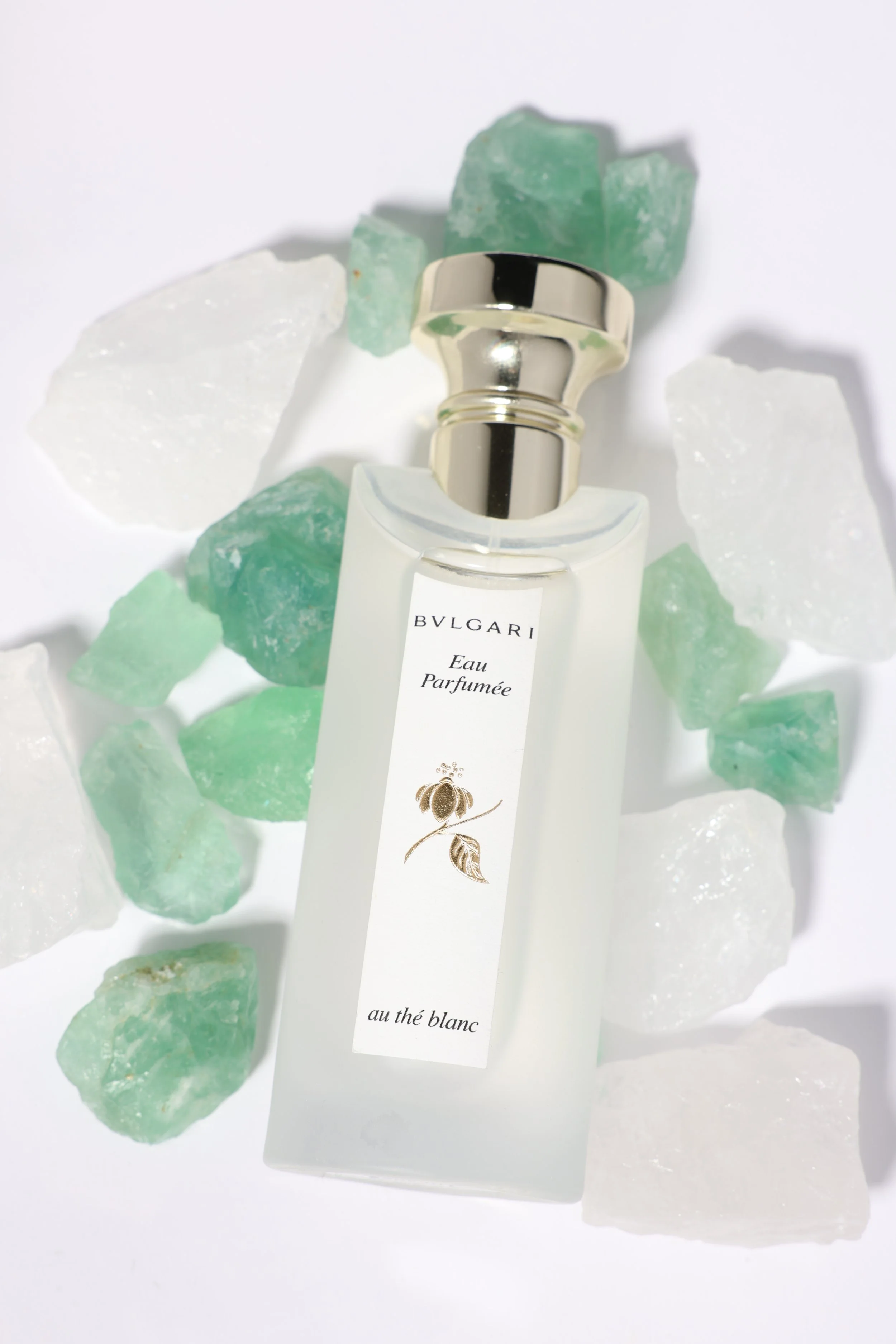 A perfume bottle labeled BVLGARI Eau Parfumée au thé blanc surrounded by white and light green rocky stones on a white background.