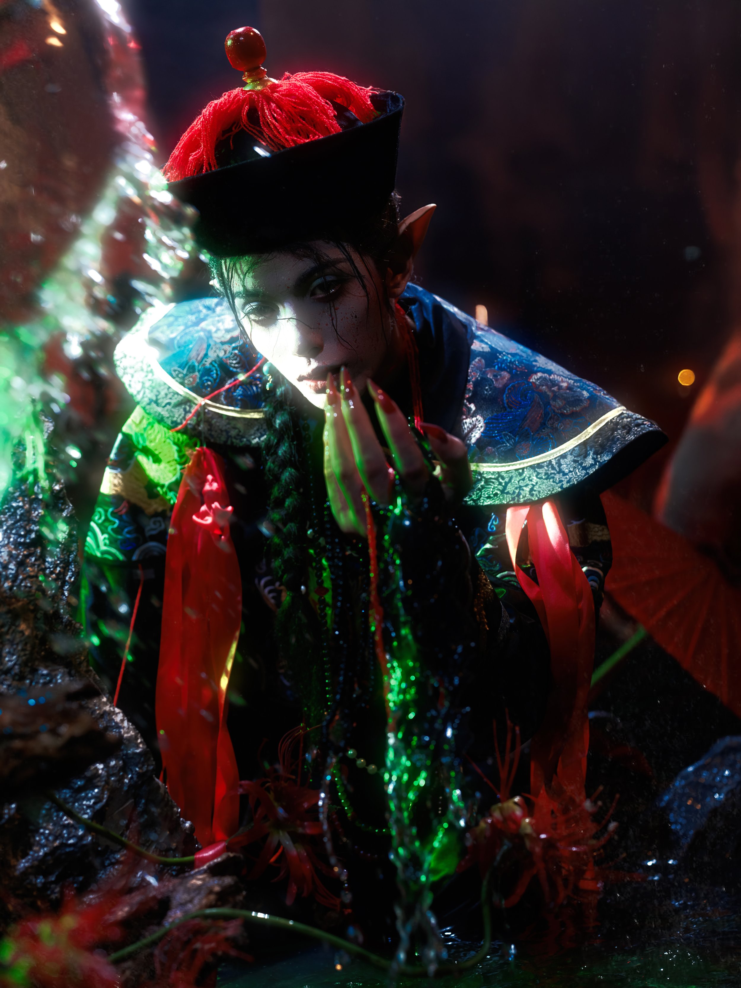 A person dressed as a fantasy character with elf-like ears, wearing an intricate, colorful costume, and a tall hat decorated with red yarn and a red ornament, kneeling near a mossy rock with glowing string lights and red flowers, illuminated by vibra