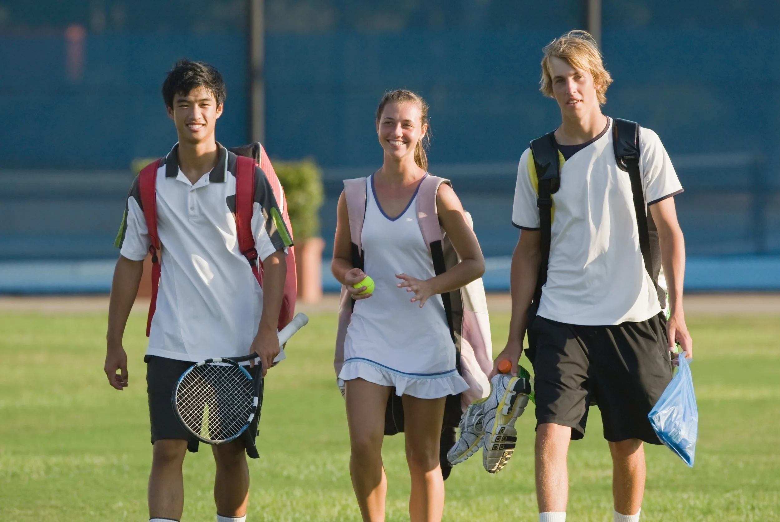Three teenagers walking on a grassy sports field carrying tennis gear and backpacks, smiling at the camera.