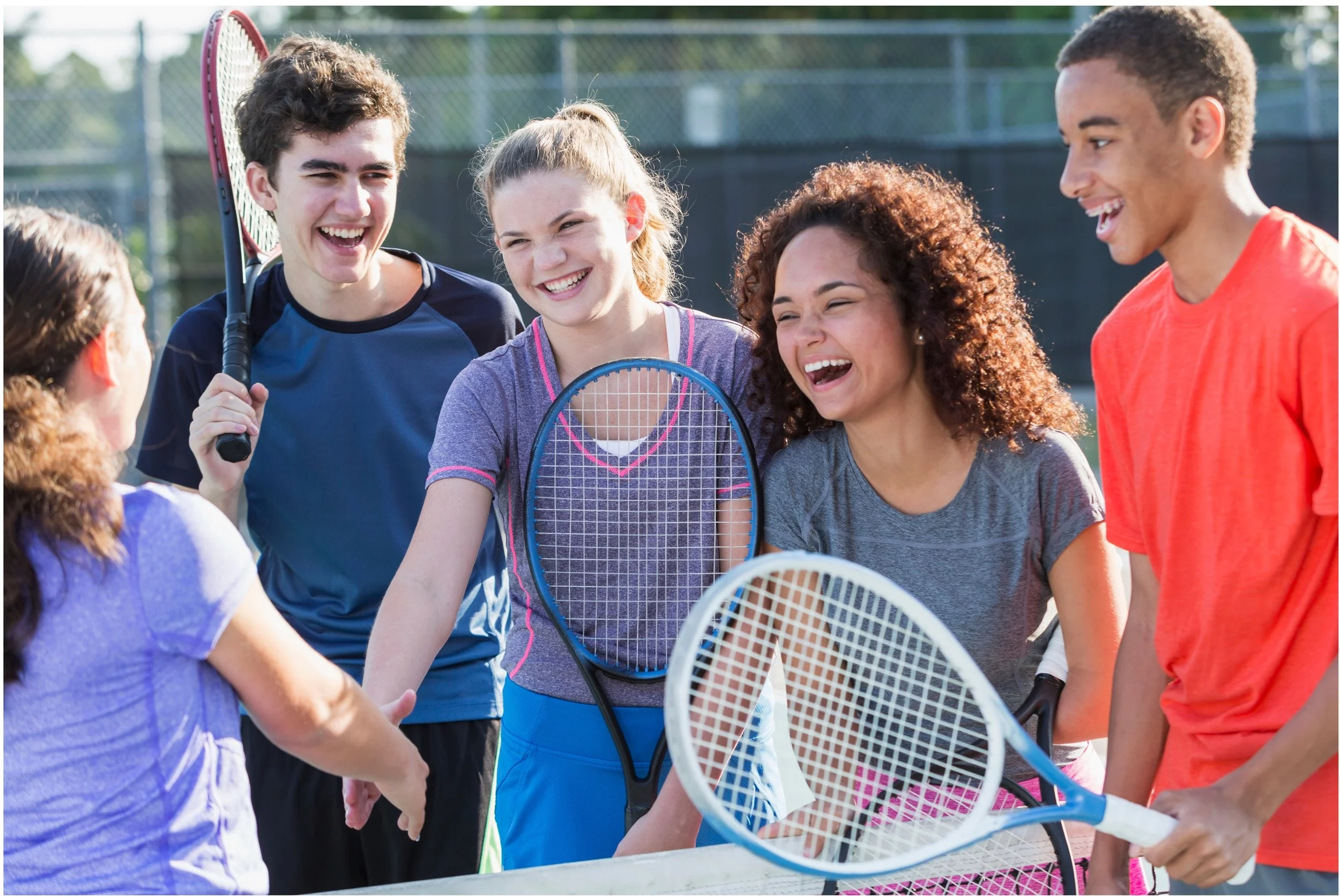 Group of five teenagers on a tennis court, smiling and talking with tennis rackets, engaging in a friendly conversation.