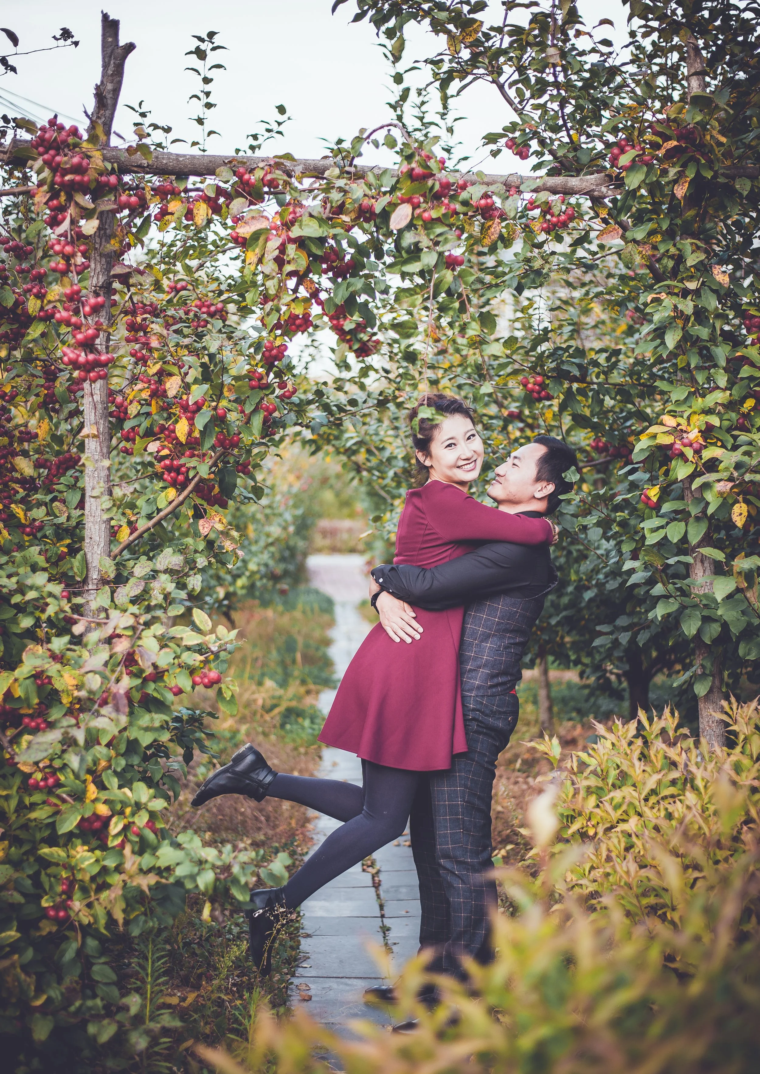 A couple happily hugging in an orchard with ripe red berries, surrounded by lush green foliage and a stone pathway.