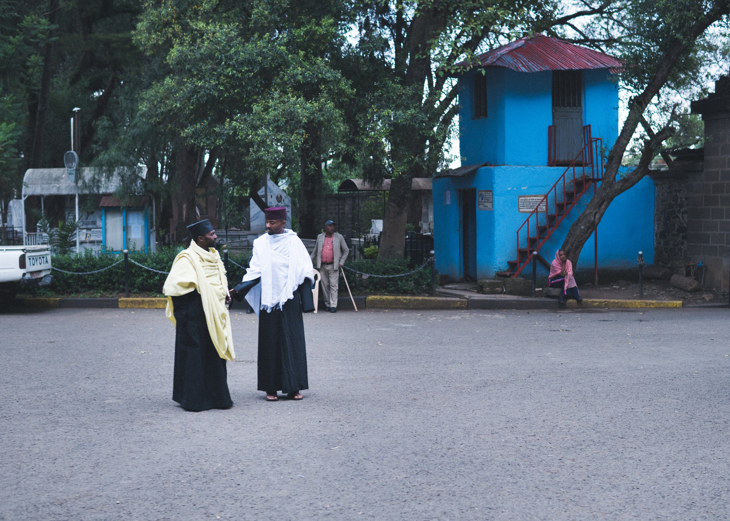 Two men in traditional Ethiopian clothing and hats conversing in an outdoor area with a blue building, trees, and a woman sitting on the curb.