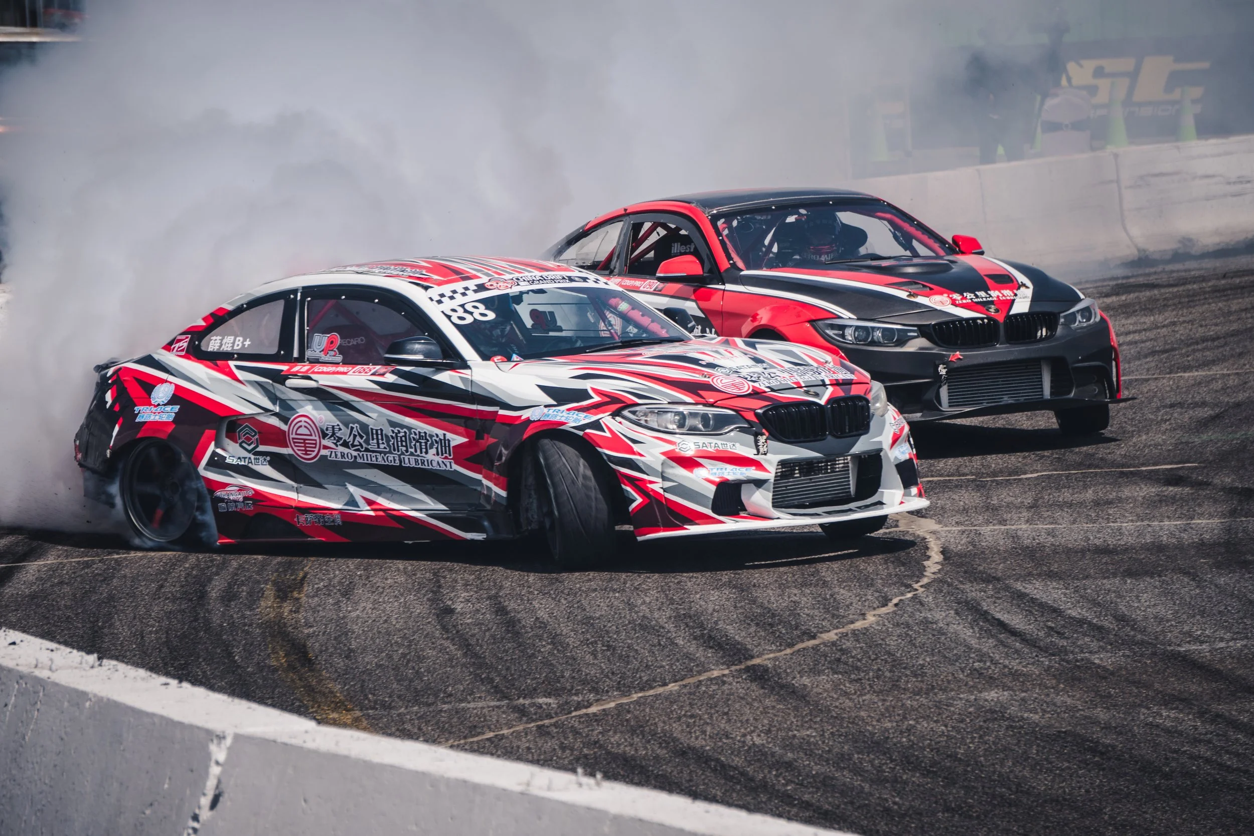 Two race cars drifting on a track, one red, black, and white with racing decals, and the other black and red, smoke billowing from the tires.